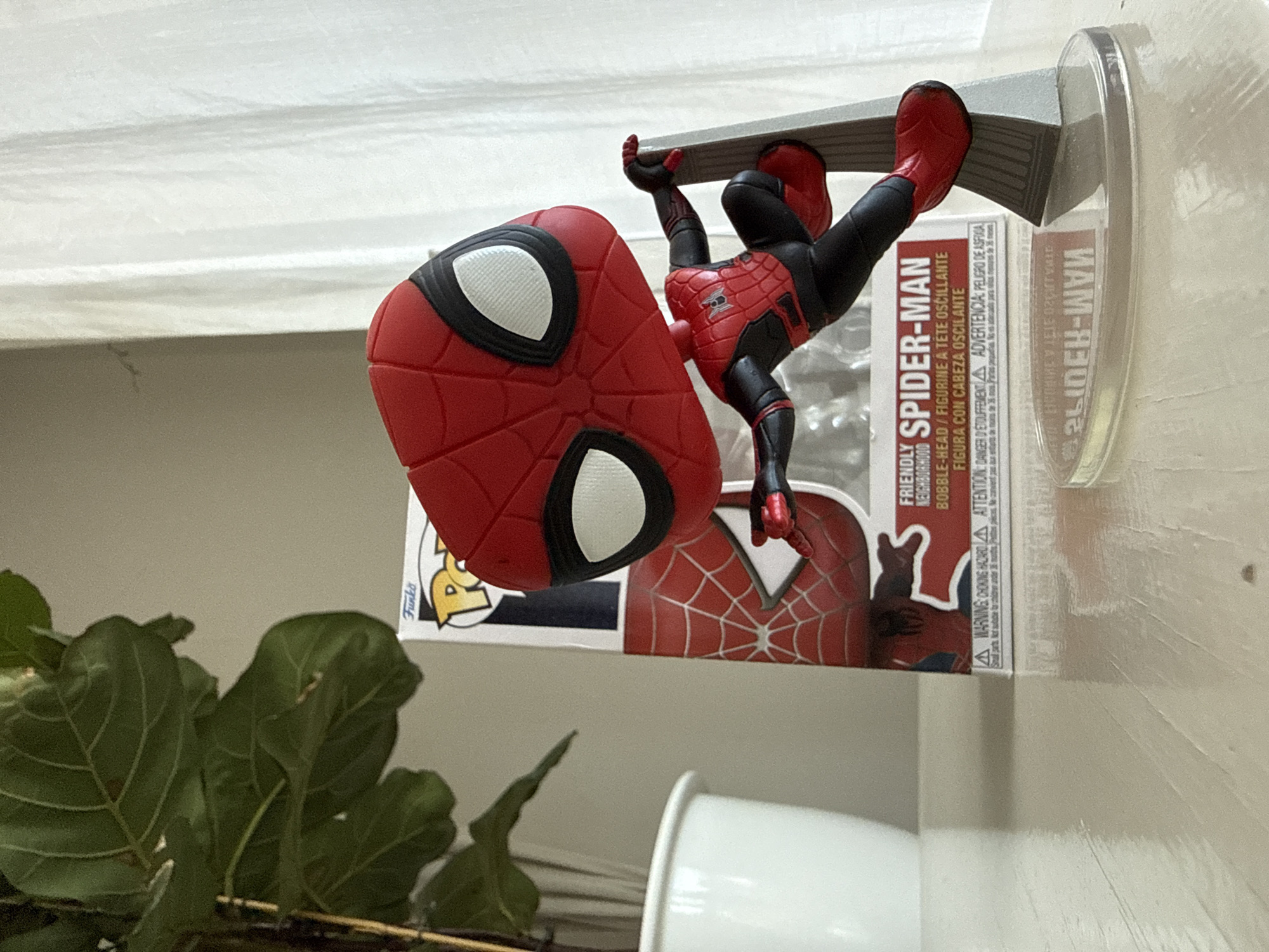 A Funko Pop bobblehead of Spider-Man is displayed on a reflective white surface, positioned to the right of a white ceramic pot containing a large green plant. The bobblehead depicts Spider-Man in his red and black suit, posed dynamically as if swinging or climbing. He is mounted on a clear plastic stand that is attached to a grey, skyscraper-like structure. Behind the bobblehead is its packaging, a Funko Pop box featuring the "Friendly Neighborhood Spider-Man" logo. The background consists of a textured grey wall on the left and a softly lit white curtain on the right. The overall scene is indoors, with the lighting suggesting it is daytime. There is no indication of the city of Bad Sachsa, Germany, in this image.