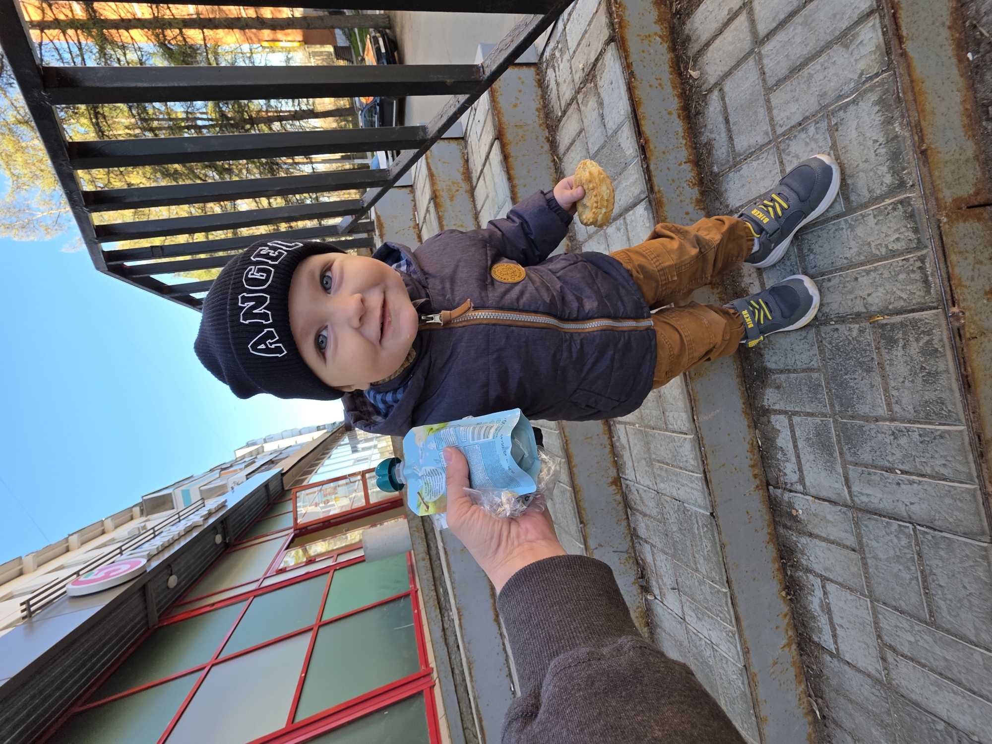 A baby wearing a black beanie with the word "ANGEL" in white lettering, a navy blue jacket with a bronze zipper and circular patch, and brown pants is lying on a concrete sidewalk. The baby is holding a fried pastry in their right hand. A person's arm is extended toward the baby, offering a pouch of baby food. To the left, the corner of a building with red and green-tinted windows is visible. Metal railings are in the background. The location is Chaikovsky, Russia.