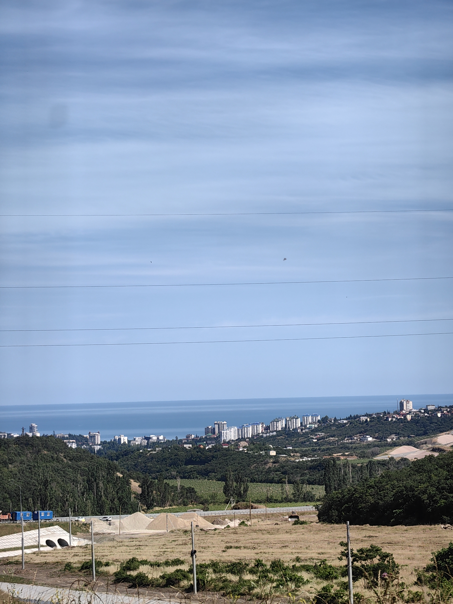 The image depicts a wide-angle view of a coastal landscape under a bright, partly cloudy sky. In the foreground, a dry, grassy field with some scattered bushes and barbed wire fencing suggests a rural or undeveloped area. Several utility poles stand in the field, one with a dark object attached. Further back, a road runs horizontally across the frame, with what appears to be construction materials, including piles of sand and what might be portable buildings, alongside it. There are also two concrete culverts visible.
Beyond the road, a lush, green hillside covered in trees slopes down towards the sea. Nestled on the hillside and along the coast is a densely populated city with numerous white, multi-story buildings, some appearing to be apartment complexes or hotels. The city stretches along the coastline, with a clear blue sea meeting the land. The sky is a pale blue with thin, horizontal clouds, indicating fair weather. The sunlight suggests it is daytime, likely late morning or early afternoon. There are no discernible people or specific activities taking place, giving the scene a sense of stillness. The location is identified as Kiparisnoe, Ukraine. The overall impression is one of a coastal city developing amidst a natural landscape.