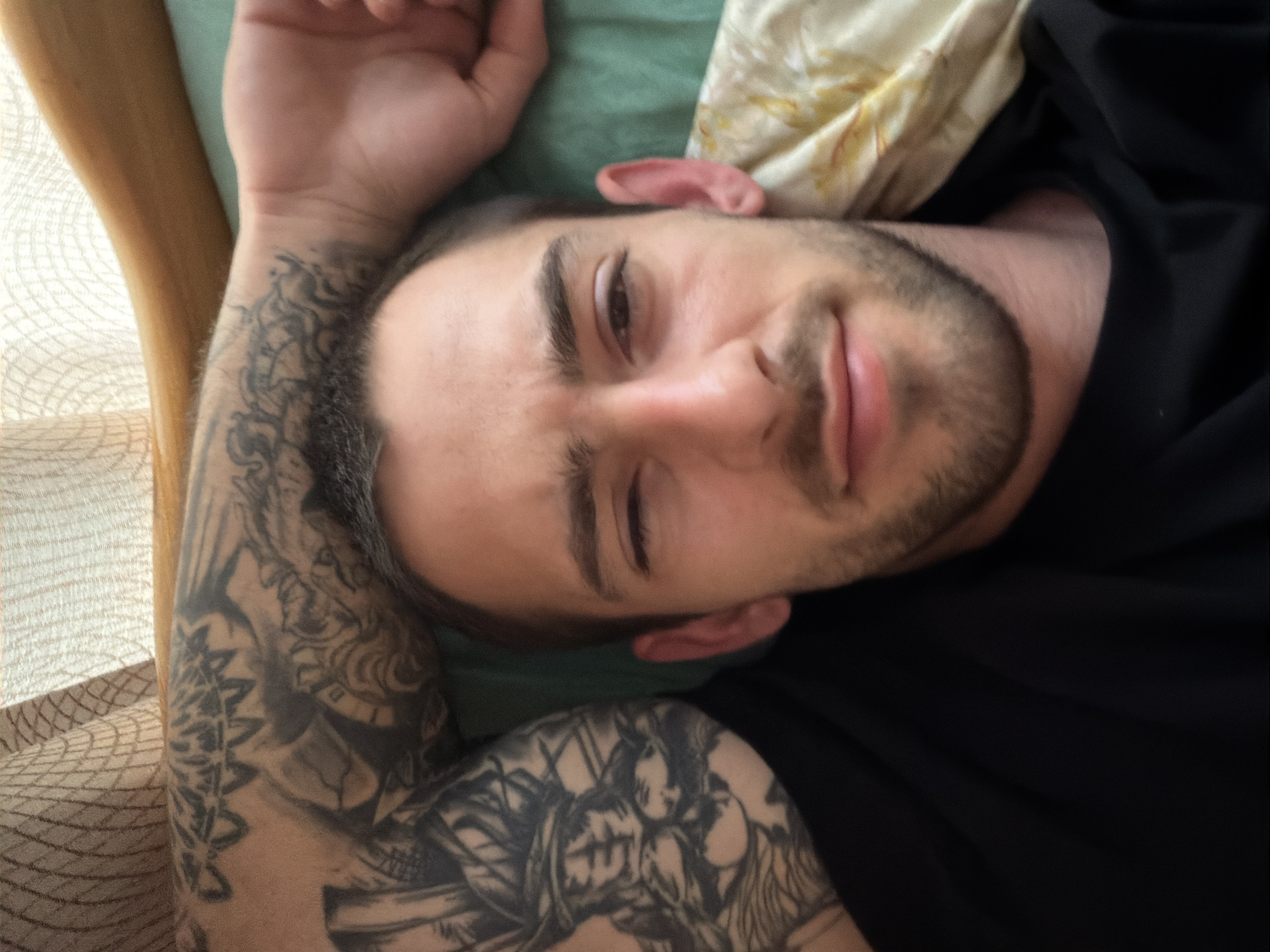 A man with a beard is lying down. His right arm, which is heavily tattooed, is raised above his head. He is wearing a black shirt. He is lying on a pillow and possibly a bed or sofa. It is not possible to determine if the photo was taken in Plovdiv, Bulgaria.