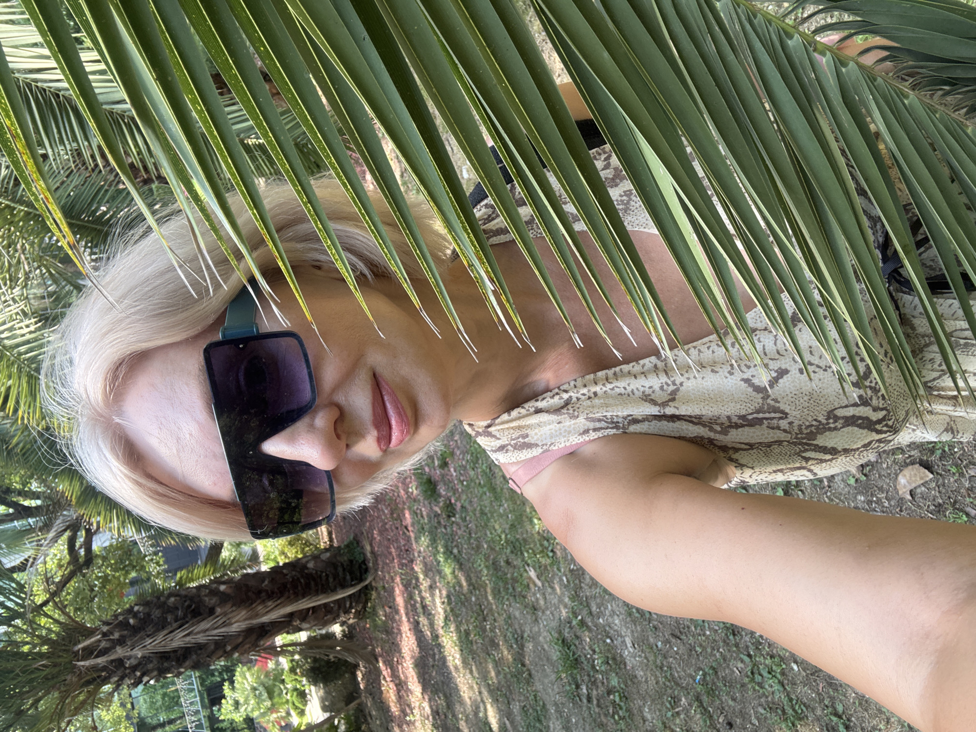 A woman with blonde hair and sunglasses is partially obscured by a large palm frond. She is wearing a dress with a snakeskin pattern and a pink bra. She appears to be outdoors in a park or garden, with palm trees and foliage in the background. The setting suggests a warm climate, possibly a resort area. The image was taken outdoors in Sochi, Russia.