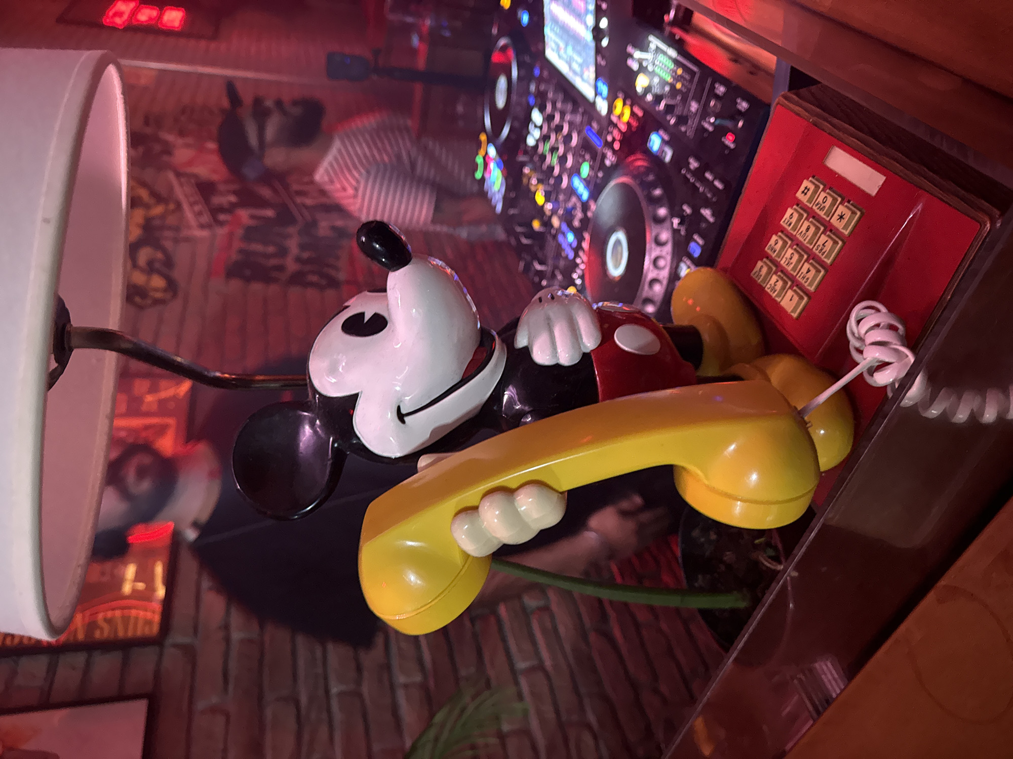 The image depicts a whimsical composition on what appears to be a reflective surface, possibly a glass table. A Mickey Mouse figure, holding a yellow telephone receiver, is the central subject. Beside the figure sits a retro red telephone with a push-button keypad. In the background, a DJ console is visible, suggesting a music-related setting. Reflections in the glass surface show a patterned rug or flooring and a lamp with a white shade. The overall lighting has a warm, reddish tone.
Given the peculiar and creative arrangement of objects, this image likely captures a scene from the Paradiso bar in Barcelona, Spain, known for its eccentric decor.