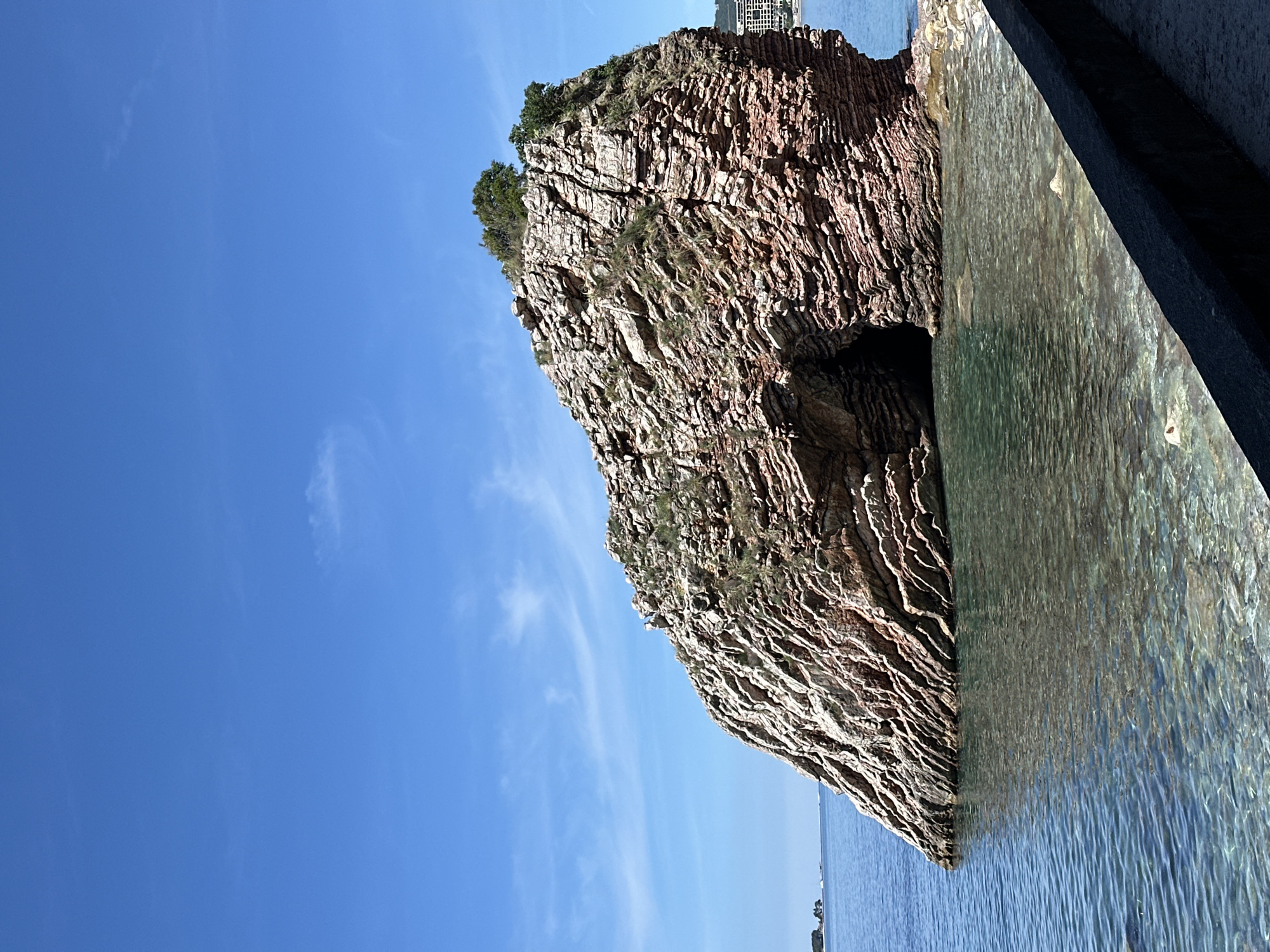 The image features a rugged, rocky islet situated in clear blue water, likely the Adriatic Sea. The islet exhibits distinct layers of sedimentary rock, with a small patch of vegetation clinging to its summit. A cave or eroded opening is visible near the base of the rock formation. A flat, man-made structure is partially visible on the right edge of the image, suggesting the presence of a pier or walkway. The clear sky and calm water indicate fair weather conditions. The presence of the islet in the sea places this scene in a coastal environment. The faint outline of buildings visible in the distance suggests that the city of Boreti is in the background.