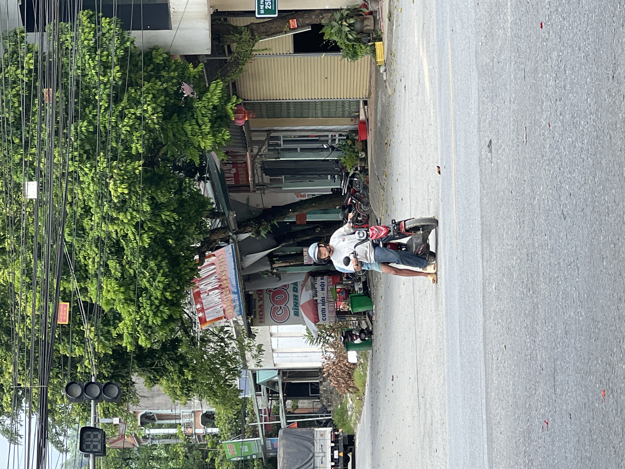 An individual wearing a white helmet, grey long-sleeved shirt, and blue jeans rides a red electric scooter down a street in Tân Quang Commune, Vietnam. The scooter has a black seat and front wheel. The individual's legs are visible, wearing light-colored sandals. The street appears to be paved with asphalt and has a yellow line dividing the lanes.
To the right of the scooter, a sign with "25B" is visible on a pole. In the background, several shops and buildings line the street. One shop has a large red and white sign with Vietnamese text. Another building has a white facade with green accents and large metal roll-up doors. Trees with lush green foliage overhang the street and buildings.
To the left of the scooter, a traffic light is visible, with three circular lights arranged vertically, and a black digital display to its left. A white truck with a grey tarp covering its load is parked on the left side of the street. Several motorcycles are parked in front of the shops. Two green trash bins are situated near a small red and white umbrella. The overall scene depicts a typical street view in a Vietnamese commune.