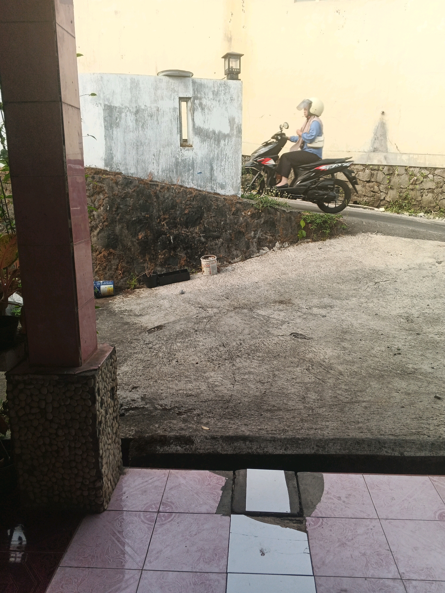 The image depicts a scene in front of a building, possibly a house, in Keji, Indonesia. In the foreground, a patterned tiled floor leads to a concrete path. To the left, a tall pillar with a stone base partially obscures the view. Beyond the concrete path, a sloping area covered in rocks and vegetation leads to a road. On the road, a person wearing a helmet and a light blue shirt rides a black motorcycle. The motorcycle has red accents on its side. Behind the motorcycle, a textured light yellow wall rises, featuring a small, rectangular window. The overall environment appears to be an outdoor residential or semi-commercial area, with a mix of natural elements and man-made structures.
