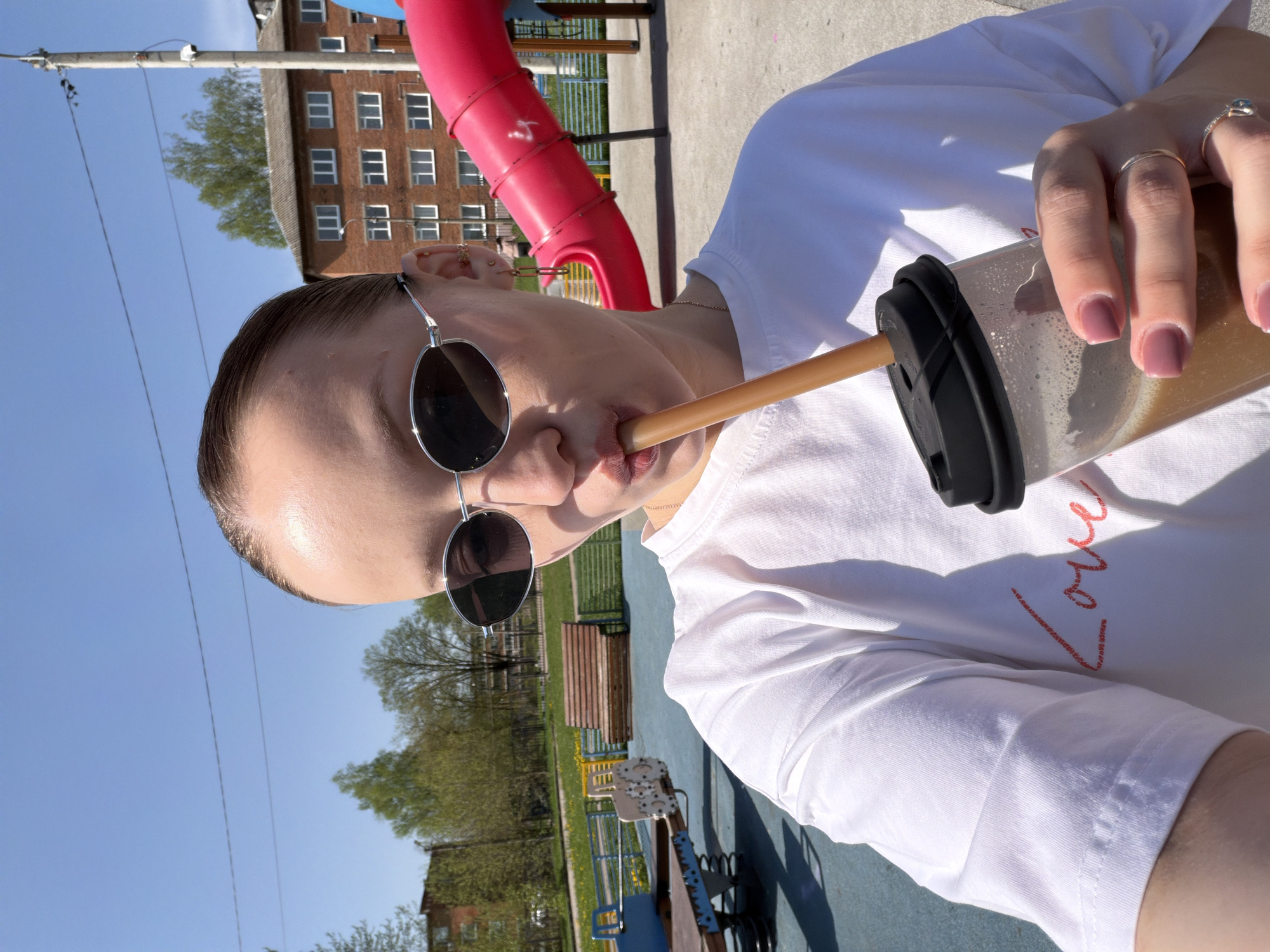 A young woman is drinking from a brown beverage container with a black lid and straw in an outdoor playground in Reshetnikovo, Russia. She is wearing round sunglasses, a white t-shirt with "Love" written in red, and has multiple rings on her fingers. The playground has a red slide and various play structures. A multi-story brick building is visible in the background, along with trees and a blue sky.