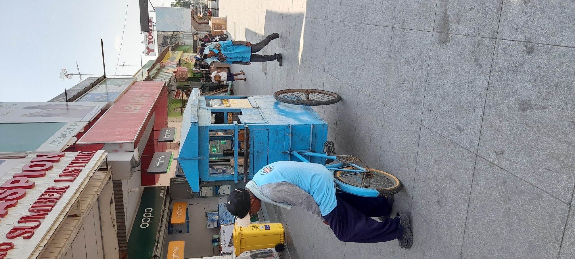 The image depicts a street scene in Pemalang, Indonesia, likely a commercial area. In the foreground, a man wearing a light blue long-sleeved shirt, dark pants, and a black baseball cap is seated on a blue, three-wheeled cart that resembles a food or beverage vendor's stall. The cart is attached to a bicycle frame, with one wheel visible in the foreground and another at the rear. The man is facing away from the camera, looking towards a storefront.
Behind the man and his cart, a sidewalk runs parallel to a row of businesses. Storefronts with signs for "SUMBER LISTRIK," "OPPO," "realme," and "Lenovo" are visible. One of the signs displays "JOERAGAN HP." The environment appears to be a sunny day, with clear skies and ample sunlight casting shadows on the pavement.
Further down the street, another individual, also wearing a light blue shirt and dark pants, is walking away from the camera. This person is wearing sunglasses and a head covering. In the background, other pedestrians and a motorcycle are visible, suggesting an active urban environment. The architecture consists of multi-story buildings with awnings and signage. A yellow trash bin is positioned near the storefronts. The ground is paved with gray tiles.