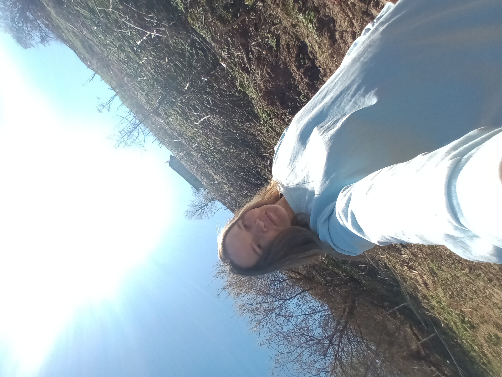 A woman is taking a selfie outside in what appears to be a sunny, outdoor environment. The background consists of foliage, trees, and blue sky. The location is Ольховка, Russia.