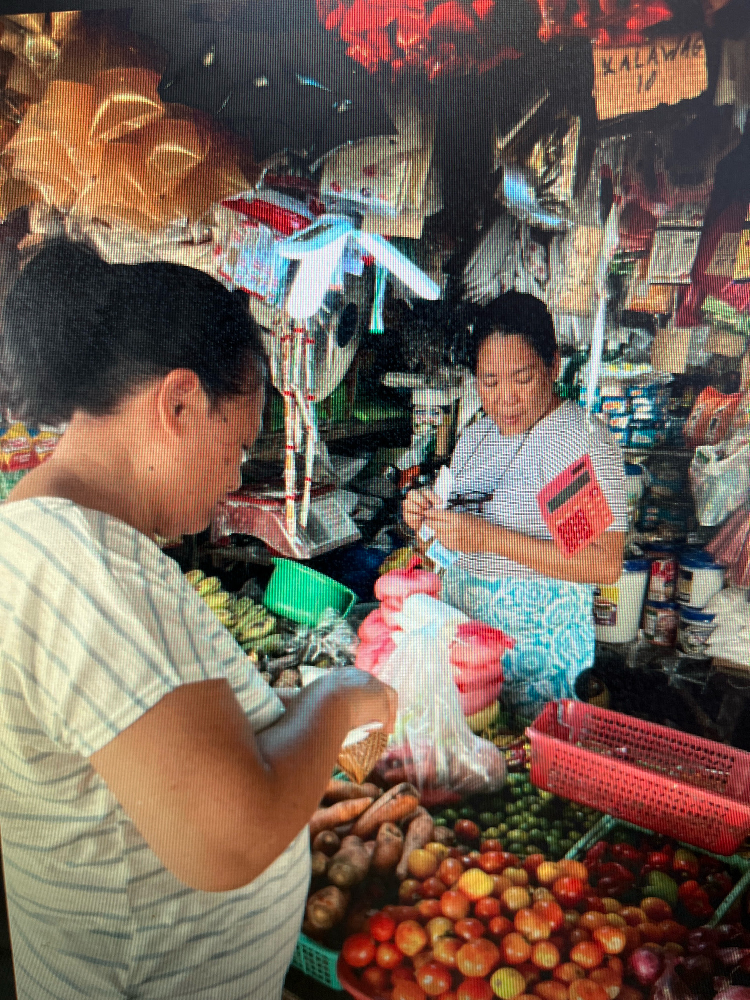 In Sejahtera, Indonesia, two women are engaged in a transaction at a market stall. The woman on the left, facing away from the camera, wears a striped shirt and is handling items from a basket. The woman on the right, facing the camera and wearing a striped top and patterned skirt, holds a calculator and appears to be ringing up the sale. The stall is laden with a variety of produce, including bananas, carrots, tomatoes, and small green fruits, along with packaged goods in the background. A red plastic basket sits prominently in the foreground, filled with more produce. The overall environment suggests a bustling marketplace.