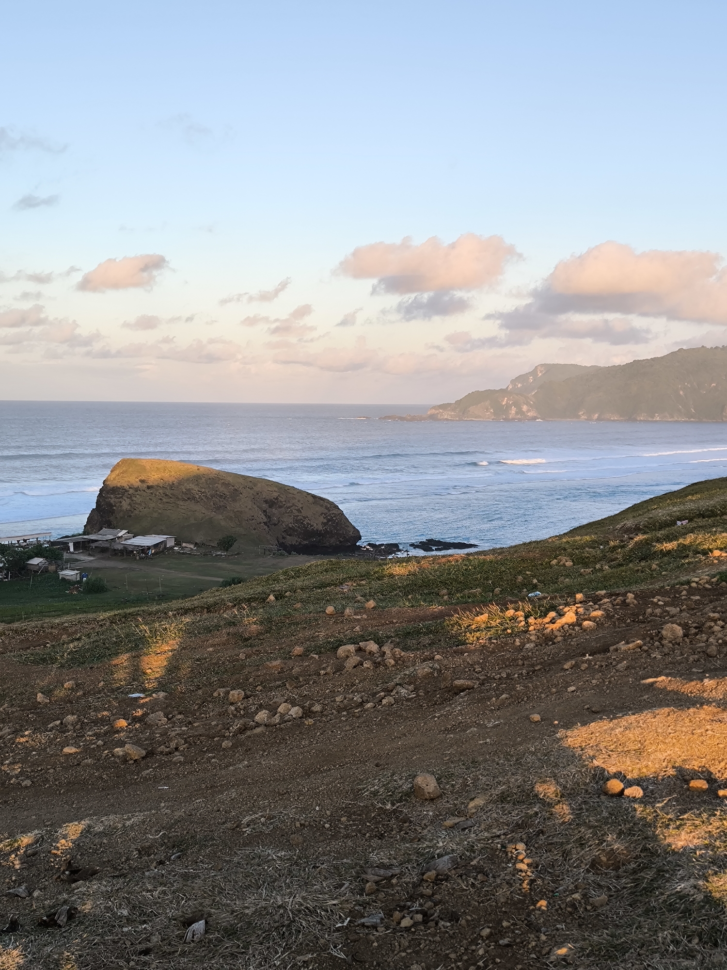 A picturesque coastal landscape is captured during either sunrise or sunset, bathing the scene in warm, soft light. The image, taken from a slightly elevated vantage point, looks out over an expansive ocean and a rugged coastline, possibly in Kuta, Indonesia.
In the foreground, a sun-kissed, rocky hillside slope covered with sparse dry grass and scattered stones leads down towards the sea. Long shadows stretch across the ground, indicating the low angle of the sun. The midground is dominated by the vast, calm ocean, where gentle waves can be seen breaking on the shore. A prominent feature is a large, dark, rounded rock formation, possibly a small island or a headland, located on the left side, close to the water's edge. Its upper surface is vibrant green and illuminated brightly by the golden light.
Nestled at the base of this large rock are several small, simple structures with corrugated metal roofs, appearing to be dwellings or shelters, suggesting a small, perhaps remote, coastal community. Further in the background, the coastline continues with distant, hazy green hills or mountains rising from the sea, adding depth to the scene. The sky above is a gradient of clear, pale blue at the top, transitioning to lighter hues near the horizon, dotted with scattered, soft, pinkish-orange clouds that reflect the early or late hour.
The overall impression is one of natural beauty and tranquility, devoid of visible people or specific activities, focusing instead on the serene interaction of land, sea, and sky at a magical time of day. No visible text is present.