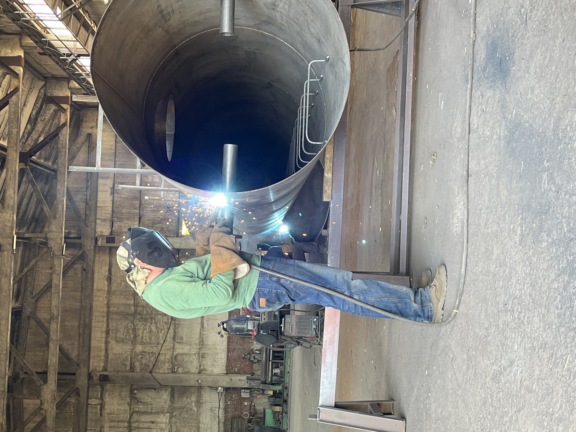 A welder, wearing a green long-sleeved shirt, blue jeans, a welding helmet, and brown work gloves, is welding a large cylindrical metal object supported by metal stands in a spacious industrial building. The welder is standing on the left side of the cylindrical object, with sparks flying from the welding torch. The cylindrical object is open at both ends, with interior metal rungs visible inside one end. In the background, other industrial equipment, including a green machine and a welding machine, are present. The floor is concrete, and the walls and ceiling appear to be made of concrete and metal. The scene depicts an industrial setting, likely a workshop or manufacturing facility.