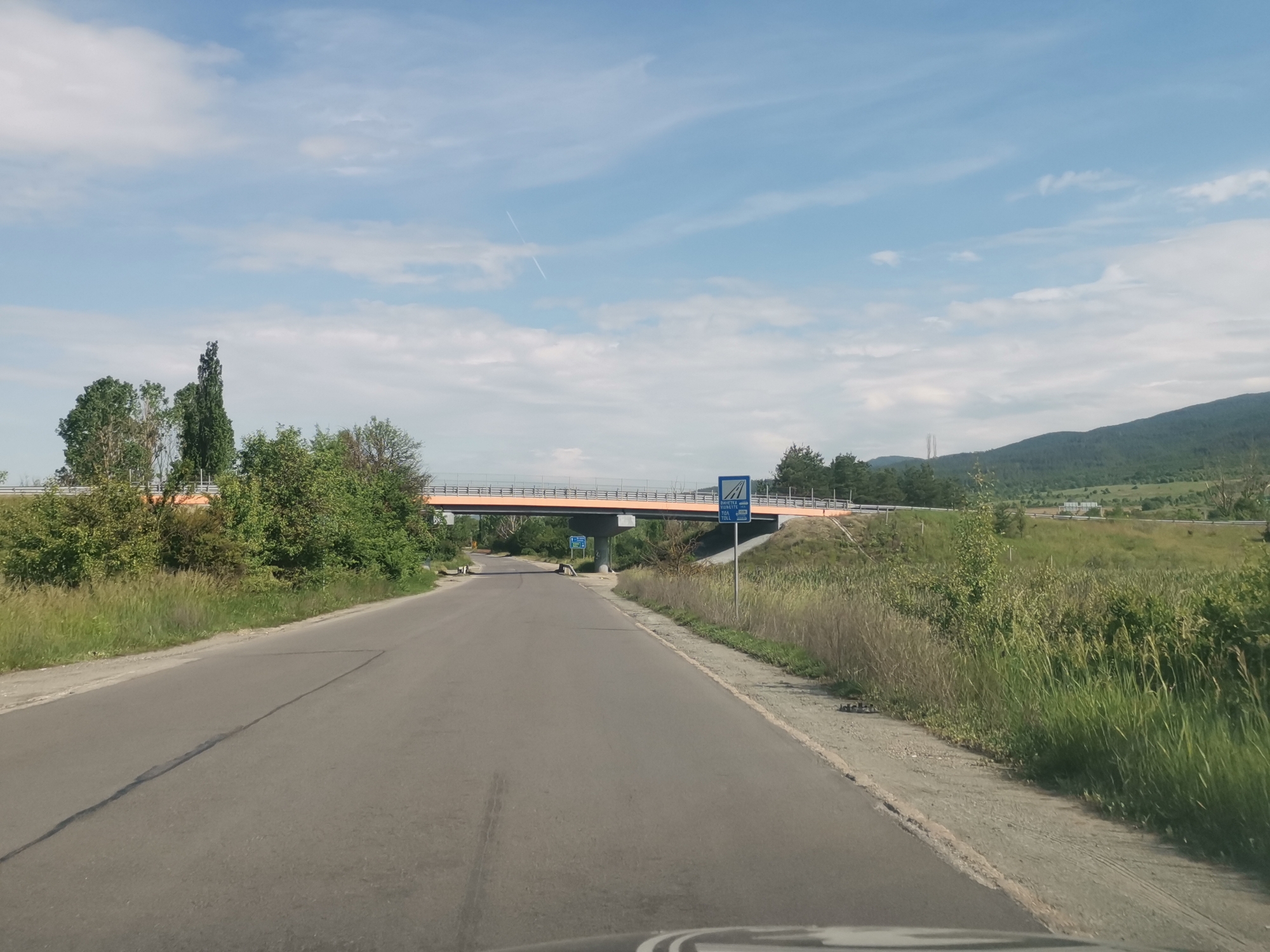 The image depicts a paved road leading toward an overpass. The overpass is painted orange and has two blue signs. One sign reads "TOLL" with other wording in a different language. The road is flanked by green vegetation and tall grass. In the background is a mountain range under a partly cloudy sky. The image appears to have been taken from a vehicle, as a portion of a dashboard is visible at the bottom of the frame. The location is near Mirovo, Bulgaria.