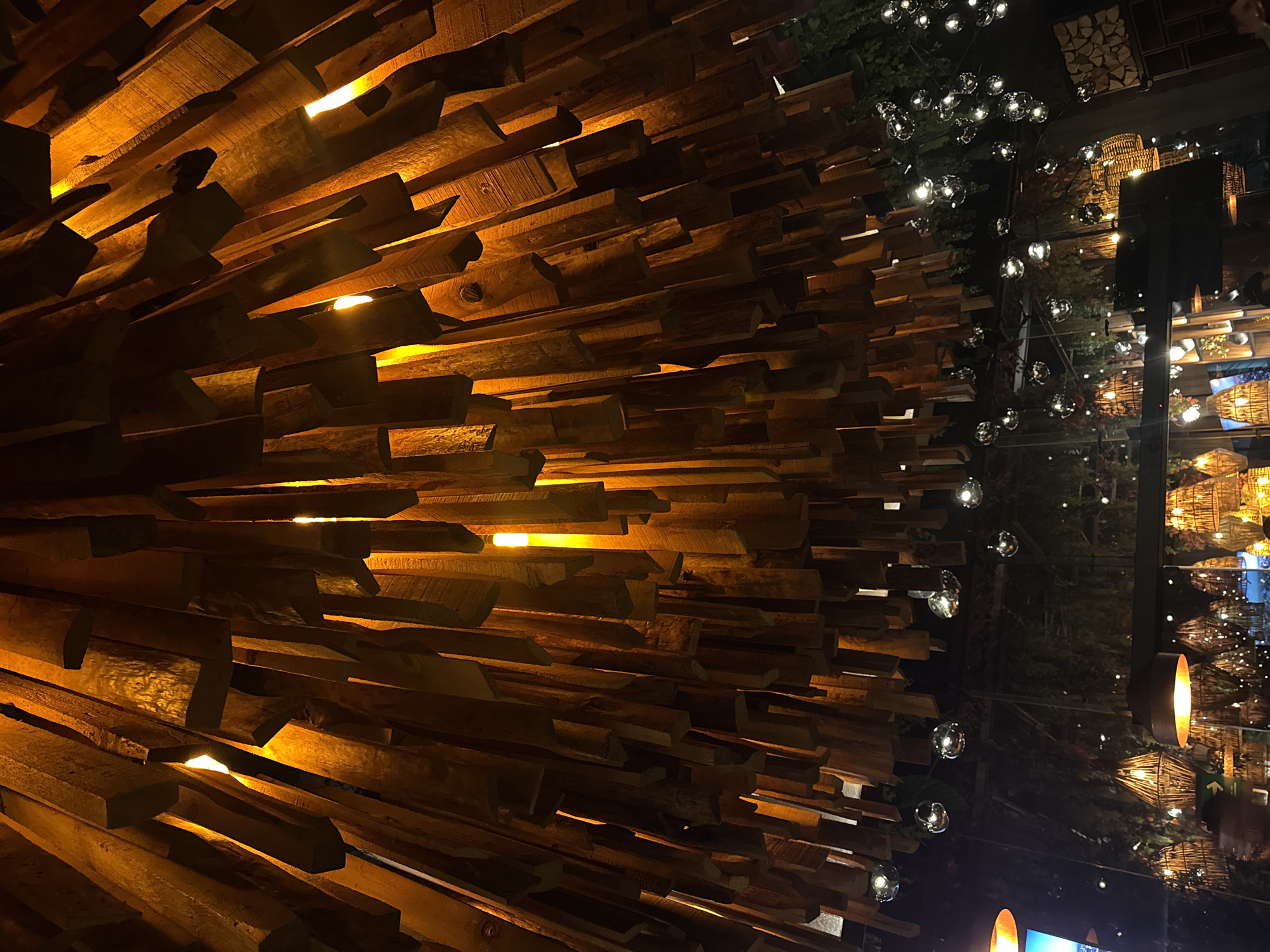 The image depicts an interior scene in what appears to be a restaurant or bar, possibly in San Bernardino Tlaxcalancingo, Mexico. A prominent feature is an elaborate ceiling installation made of irregular, stacked wooden planks illuminated with warm, orange light emanating from in between them. The right side of the image shows a reflection of the bar's interior, including decorative lights hanging from the ceiling and wicker-like baskets. A partial view of outdoor vegetation is reflected in a dark pane. No people are clearly visible. The focus of the image is on the ceiling and its interplay of wood, light, and reflections.