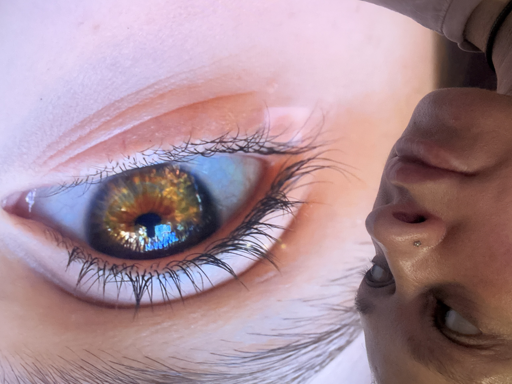 This vertically oriented image features a detailed close-up of a human eye, likely displayed on a digital screen due to subtle pixelation. The prominent iris is a vibrant mix of amber, golden-brown, and copper tones, with hints of blue along its edges, and a dark, central pupil. The pupil distinctly reflects bright, structured blue and white light, suggesting a window or another screen as the light source. Long, dark eyelashes frame the eye, and the surrounding skin shows visible texture, including fine lines and pores, indicating an extreme close-up perspective.
To the right of the main eye, an inverted human face is partially visible. This face includes a nose with a small, shiny stud piercing and part of an eye looking downwards. This element appears to be a reflection of the person taking the photograph or an observer looking at the screen. The scene captures a moment of observation or interaction with a digital display. The lighting is bright and direct, contributing to the vivid reflections. No text or specific location cues are present within the image.