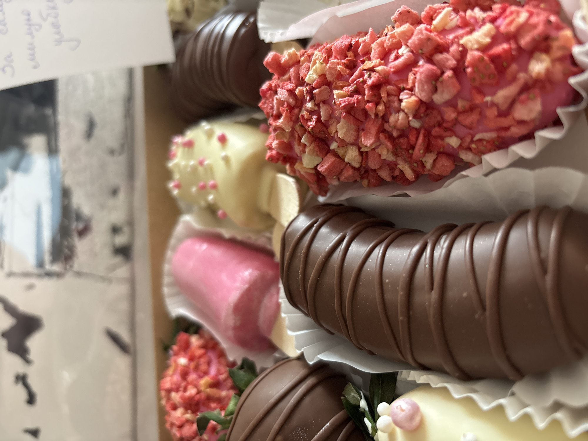 The image shows a close-up of a box filled with chocolate-covered bananas. The bananas are decorated with different toppings, including pink sprinkles, white chocolate with pink dots, and chopped strawberry pieces. Some of the bananas are covered in milk chocolate with a drizzled chocolate design. The bananas are arranged in white paper liners within the box. A small, out-of-focus card with text in an unidentified language is partially visible in the background. The location, Perm, Russia, is not discernible from the visual elements.