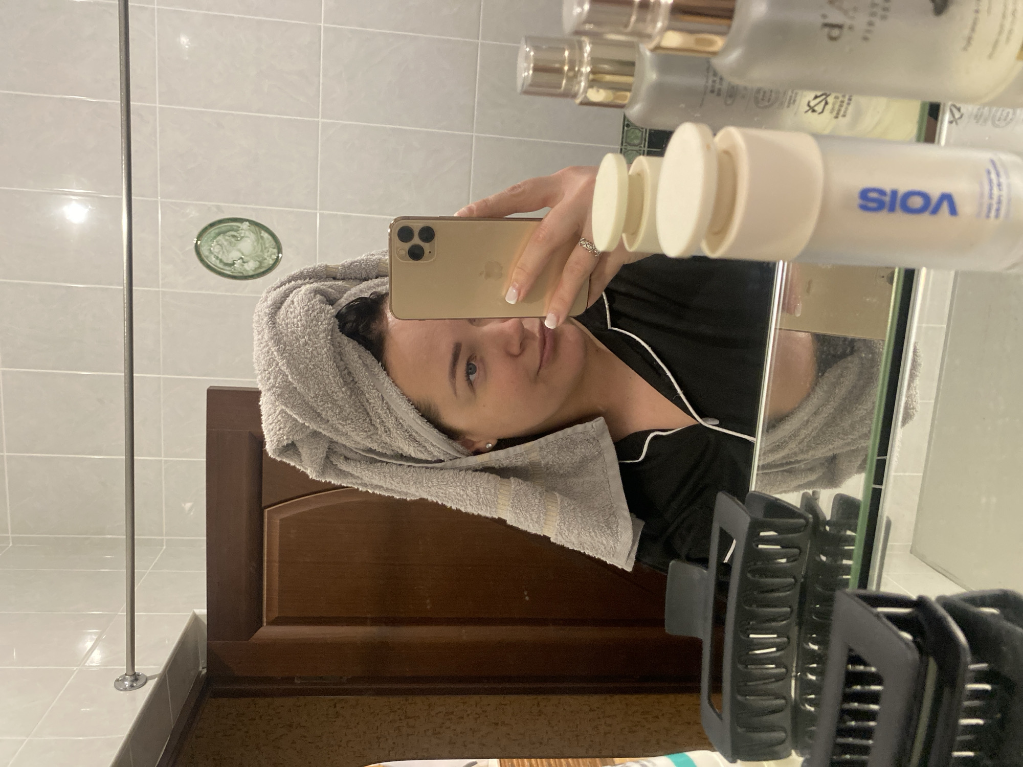 A woman with a towel wrapped around her head is taking a mirror selfie with a gold-colored iPhone. She is wearing a black top with white trim and a ring on her finger. In the mirror's reflection, various cosmetic bottles are visible. The bathroom has light-colored tiles and a small decorative cameo on the wall. A dark-colored wooden vanity and black hair clips are also in view. The setting is likely a private bathroom. The location in Sochi, Russia cannot be ascertained from the image.