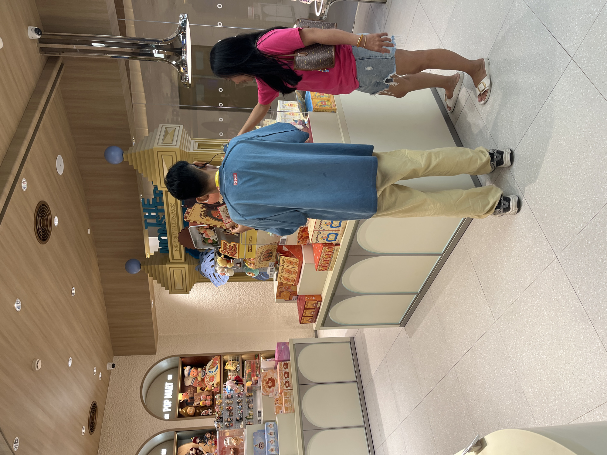 In a retail store in Kuta, Indonesia, two individuals are browsing merchandise. A person wearing a blue t-shirt with a red "POP MART" logo on the back and light yellow pants stands facing away from the camera, holding a box. To their right, a woman in a pink t-shirt and distressed denim shorts is looking at merchandise on a display. She is holding a brown handbag with a gold chain and has her right arm extended, touching a product on the shelf. The environment is a brightly lit store with tiled flooring and display shelves filled with boxes and figurines. There is a sign visible with "THE MONSTER" and "X HOW2WORK" branding.