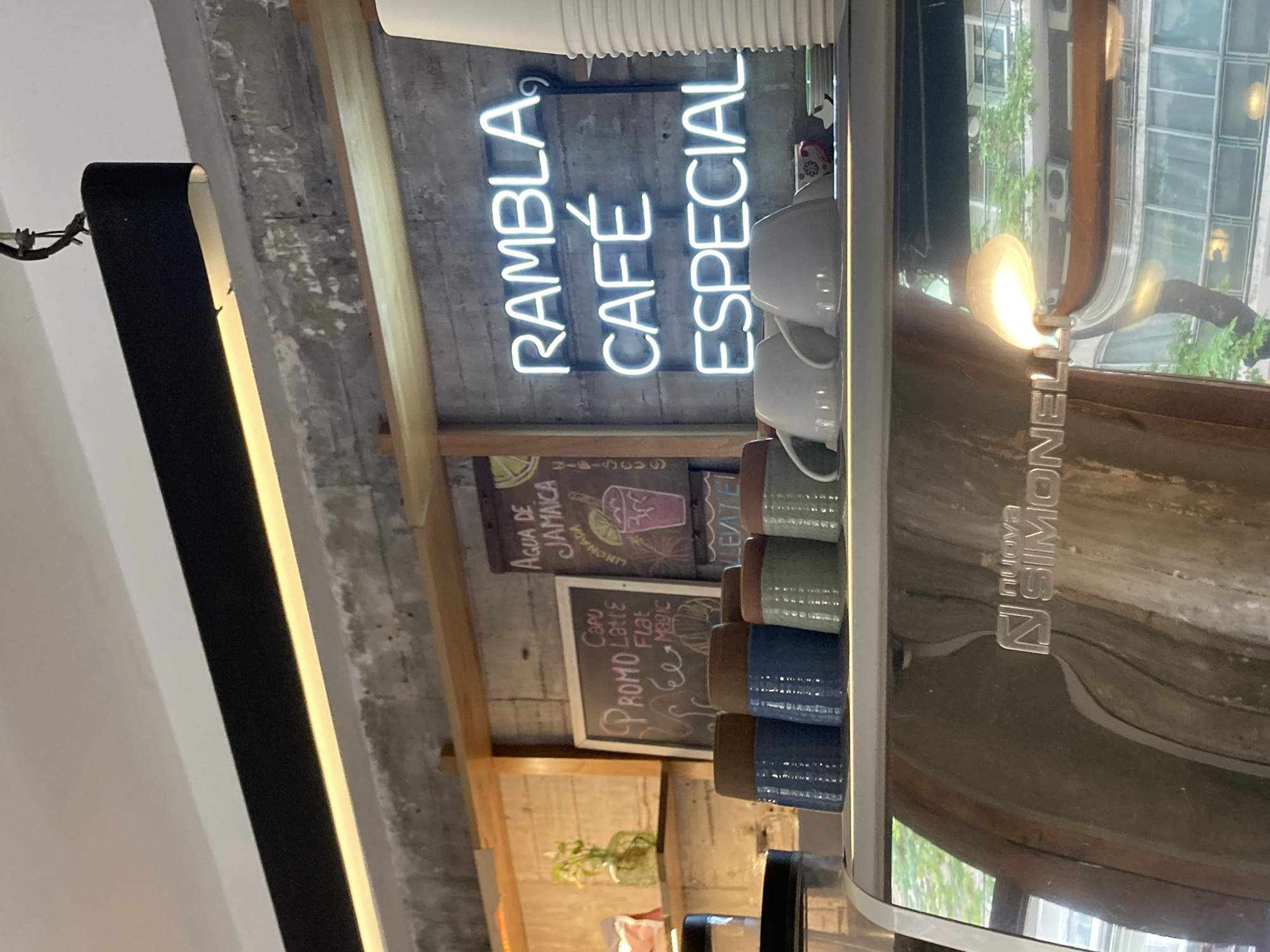 Here is a detailed description of the image:
The image shows the interior of a café. A neon sign that reads "RAMBLA CAFÉ ESPECIAL" is a central focus. Below the sign, there's a chalkboard with the words "AGUA DE JAMAICA" written on it and a small illustration of a beverage.
To the right of the sign are several stacked coffee mugs. A shiny metallic espresso machine, branded "Nuova Simonelli", is visible. The reflection on the machine suggests an outdoor urban setting with trees and buildings.
A smaller chalkboard beneath the "AGUA DE JAMAICA" one displays promotional offers like "CPROMO" and various coffee types such as "Capu", "Latte", "Flat", and "Magic". A decorative plant can be seen on a shelf below the chalkboards.
A large, rectangular black light fixture hangs on the left side of the frame, suggesting the café has modern interior design elements. The overall environment appears urban, possibly in a city location. The specific city or precise details are not discernible from the visual information.