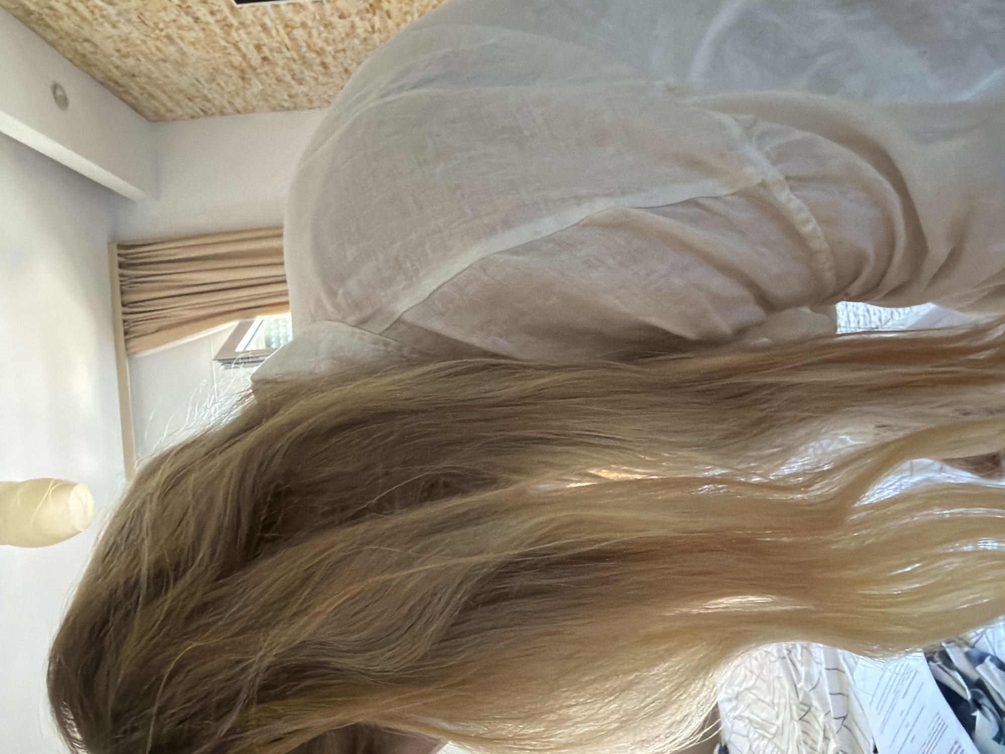 This is a close-up, low-angle shot of a person's long, blonde hair, which cascades down and fills the majority of the frame. The hair appears to be slightly tousled and is illuminated by what seems to be natural light. The person is wearing a loose-fitting, light-colored garment, possibly made of linen, which is draped over their shoulders and back.
In the background, the setting suggests an indoor environment. The ceiling is visible at the top of the image and is constructed from textured wooden panels. White walls and a window with beige-colored blinds are also apparent, indicating a room with natural light. A light fixture is visible on the wall, and a decorative element, possibly a lamp or light shade, is seen on the left side of the image. The floor area visible at the bottom of the frame shows a white, textured surface with some patterned fabric.
The overall impression is a candid shot, possibly taken in a home or accommodation, given the domestic setting. The lighting and the focus on the hair suggest an intimate or personal moment. The location context of Budva, Montenegro, provides a geographical setting for this scene, although no specific visual cues directly confirm this location. There is no visible text in the image.