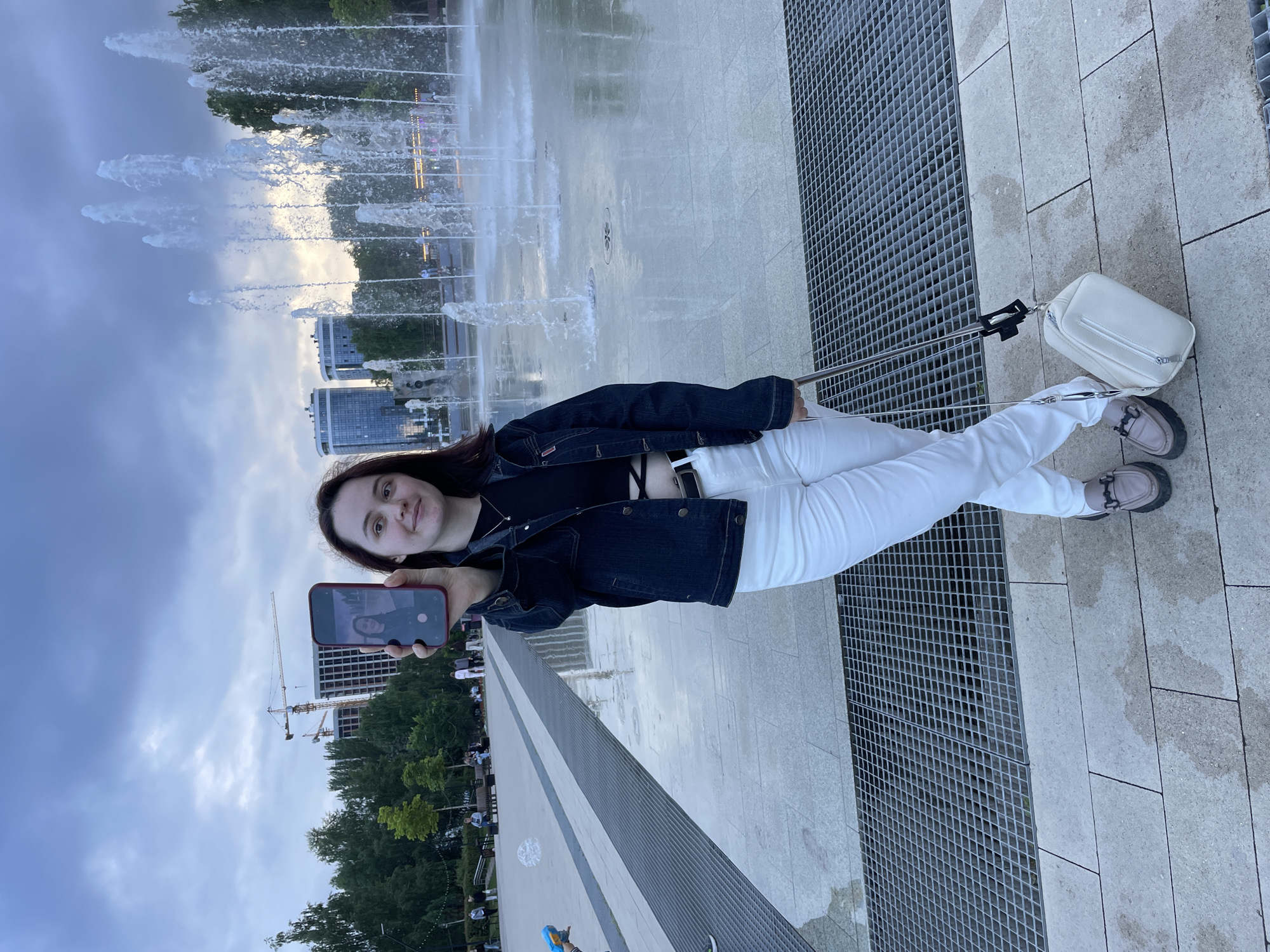 A young woman is standing in a public square, holding up a smartphone in landscape orientation to take a selfie. She is wearing a dark denim jacket, a black crop top, white pants, and light brown loafers. She has dark hair and is smiling at the camera. In the background, there is a large fountain with water jets, several modern buildings, trees, and construction cranes, suggesting the location is a developing area. The ground is paved with light-colored stone, and there are metal grates visible. The sky is overcast and appears to be dusk. The woman is also holding a white handbag attached to a metal pole, possibly a selfie stick. The overall scene depicts an urban outdoor setting.