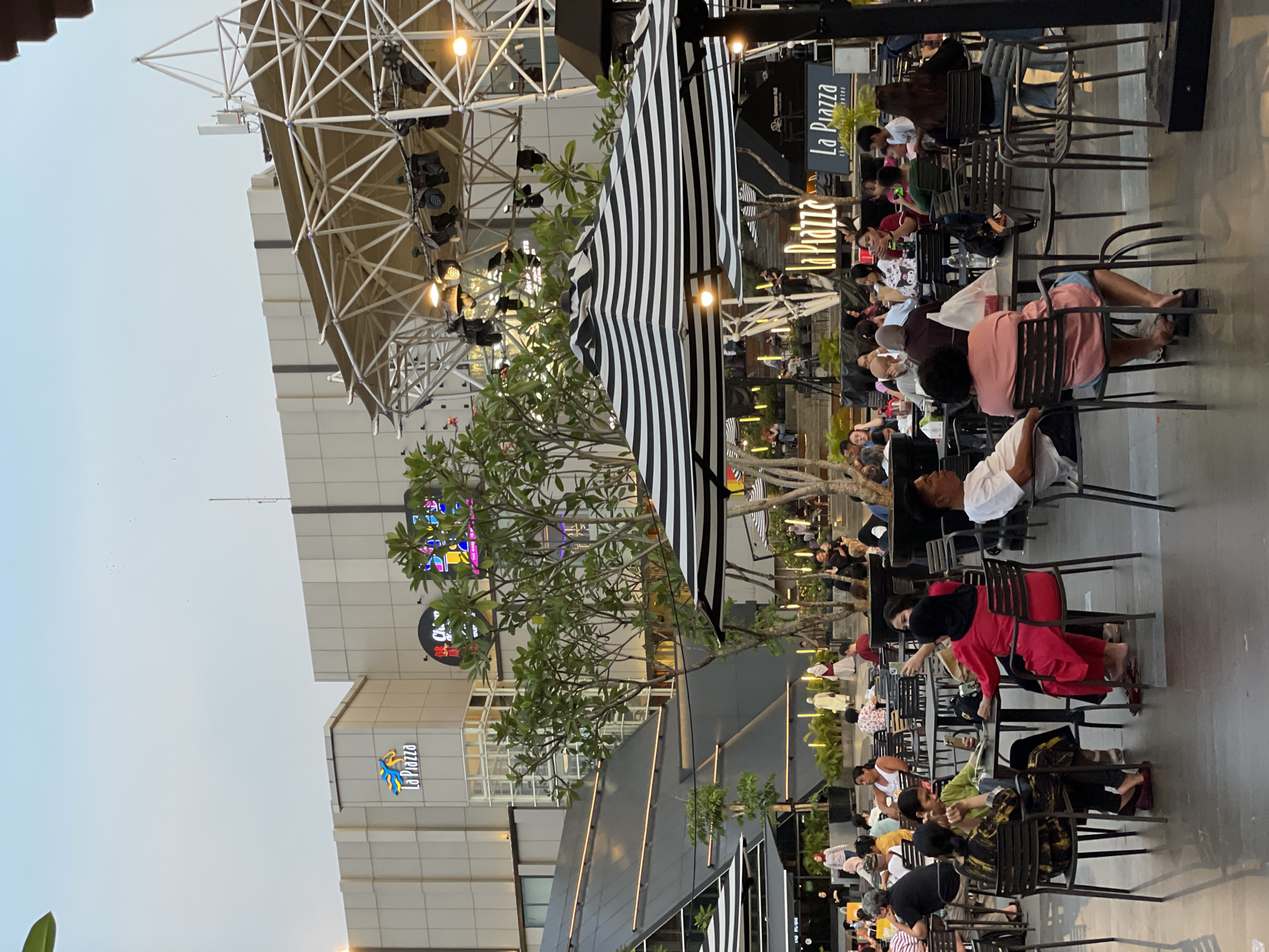 The image depicts an outdoor scene in North Jakarta, Indonesia, likely an open-air plaza area associated with a shopping mall called "La Piazza." The scene is active with numerous individuals seated at tables, engaged in dining or socializing. Black metal chairs and tables are arranged in rows. The mall's facade is visible in the background, featuring the "La Piazza" logo. A large white scaffolding structure with stage lighting stands above the seated area. A black and white striped canopy provides shade. The scene appears to be captured during late afternoon or early evening.