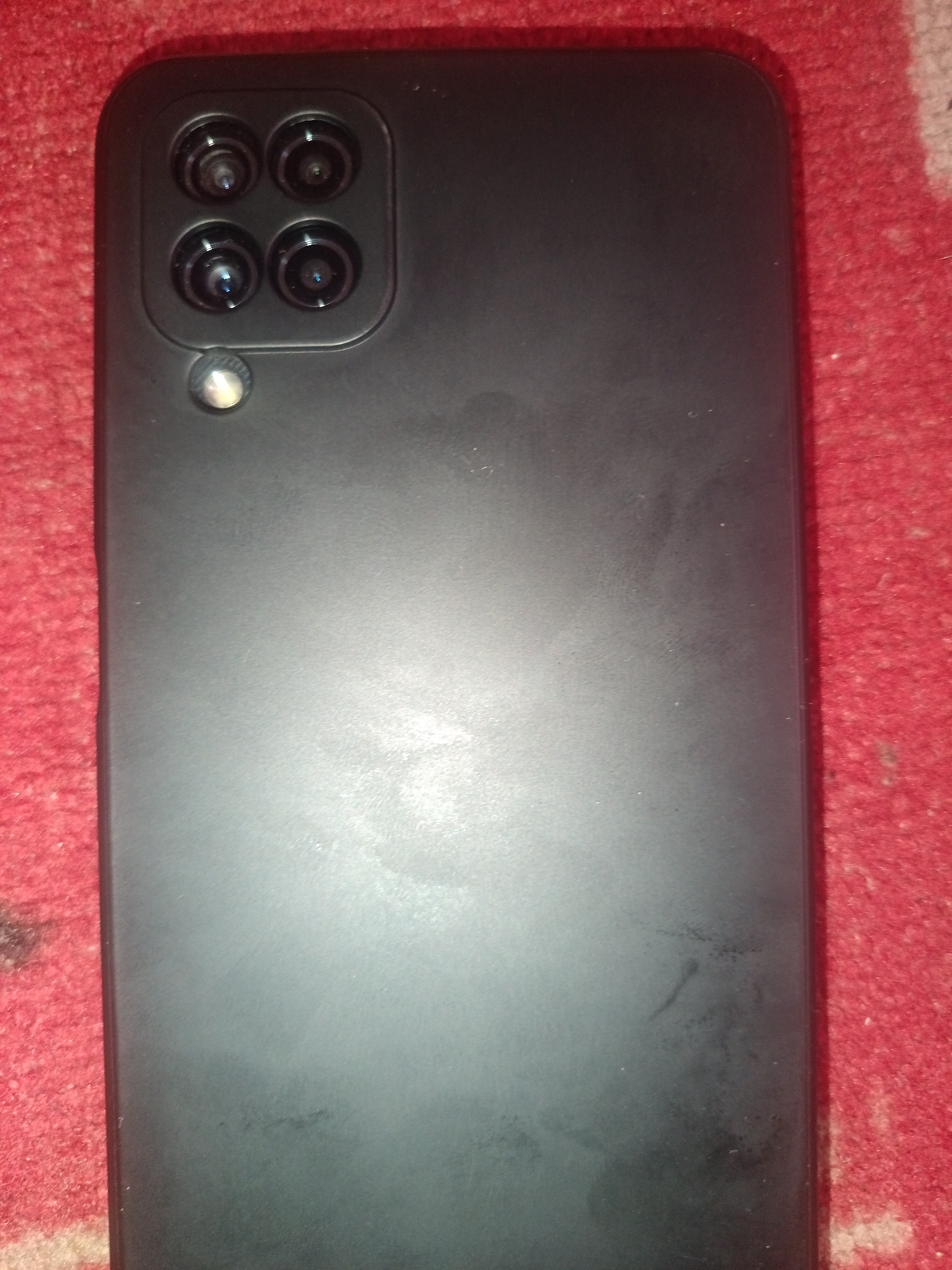 The image shows the rear of a black smartphone, likely a Samsung Galaxy A12, resting on a red, textured surface, possibly a carpet or blanket. The smartphone features a quad-camera system arranged in a square formation on the upper left side of its back. Below the cameras, a single LED flash is visible. The surface of the phone appears slightly smudged and dusty. The red background is textured with a raised pattern, consistent with a rug or similar fabric. The lighting in the image is dim, casting subtle shadows. The image provides no information about its location in Novokuznetsk, Russia, or any human presence.