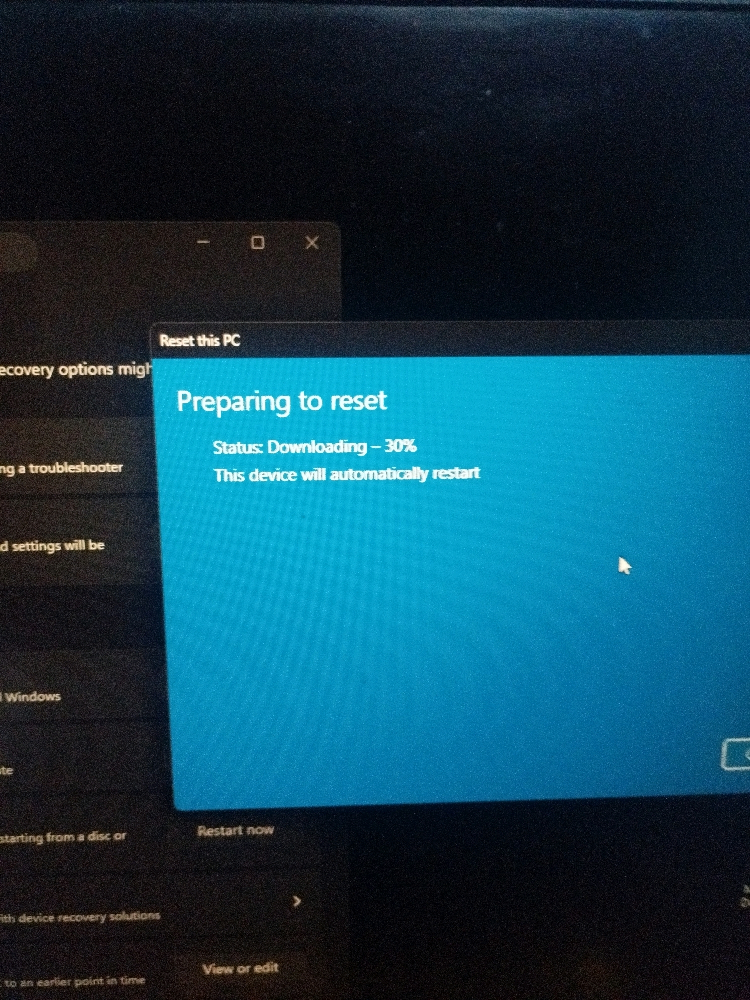 The image shows a computer screen displaying a Windows "Preparing to reset" message. A blue dialog box with the title "Preparing to reset" indicates a "Status: Downloading - 30%". Below this, it states "This device will automatically restart". In the background, another window labeled "Reset this PC" is visible, along with other options on the left side of the screen such as "recovery options might", "ng a troubleshooter", and "d settings will be". The screen appears to be from a personal computer or laptop. The image is a photograph taken of the screen, with some glare and slight blur. There is no information within the image to indicate a location in Madhyapur Thimi, Nepal.