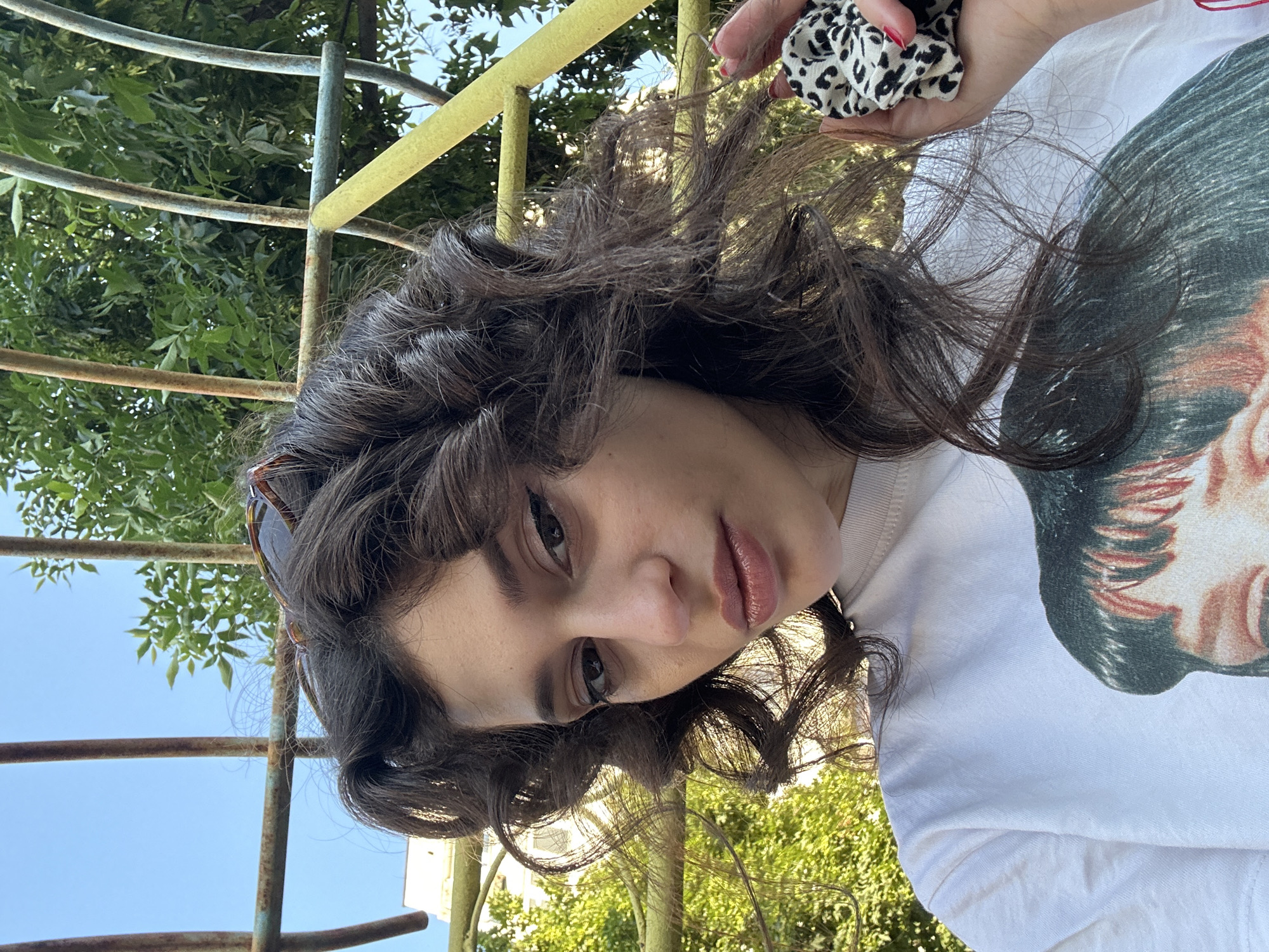 This is a close-up, vertical, outdoor, daylight photograph of a young person. The subject has dark, curly hair, with some strands falling across their face. They are wearing a white t-shirt with a graphic print of a person's face. The subject's face is turned towards the camera, with their lips slightly parted. They are wearing sunglasses on top of their head. In the upper right corner, a hand with red-painted fingernails is holding a black and white spotted scrunchie.
The background consists of greenery, likely trees, and a bright blue sky. There are also some rusted metal bars, possibly part of a playground or fence, in the foreground and to the left of the subject. The lighting suggests it is daytime, with the sun casting some shadows and highlighting the subject's hair. The overall mood is casual and relaxed. The location context is Sofia, Bulgaria. There is no visible text in the image.