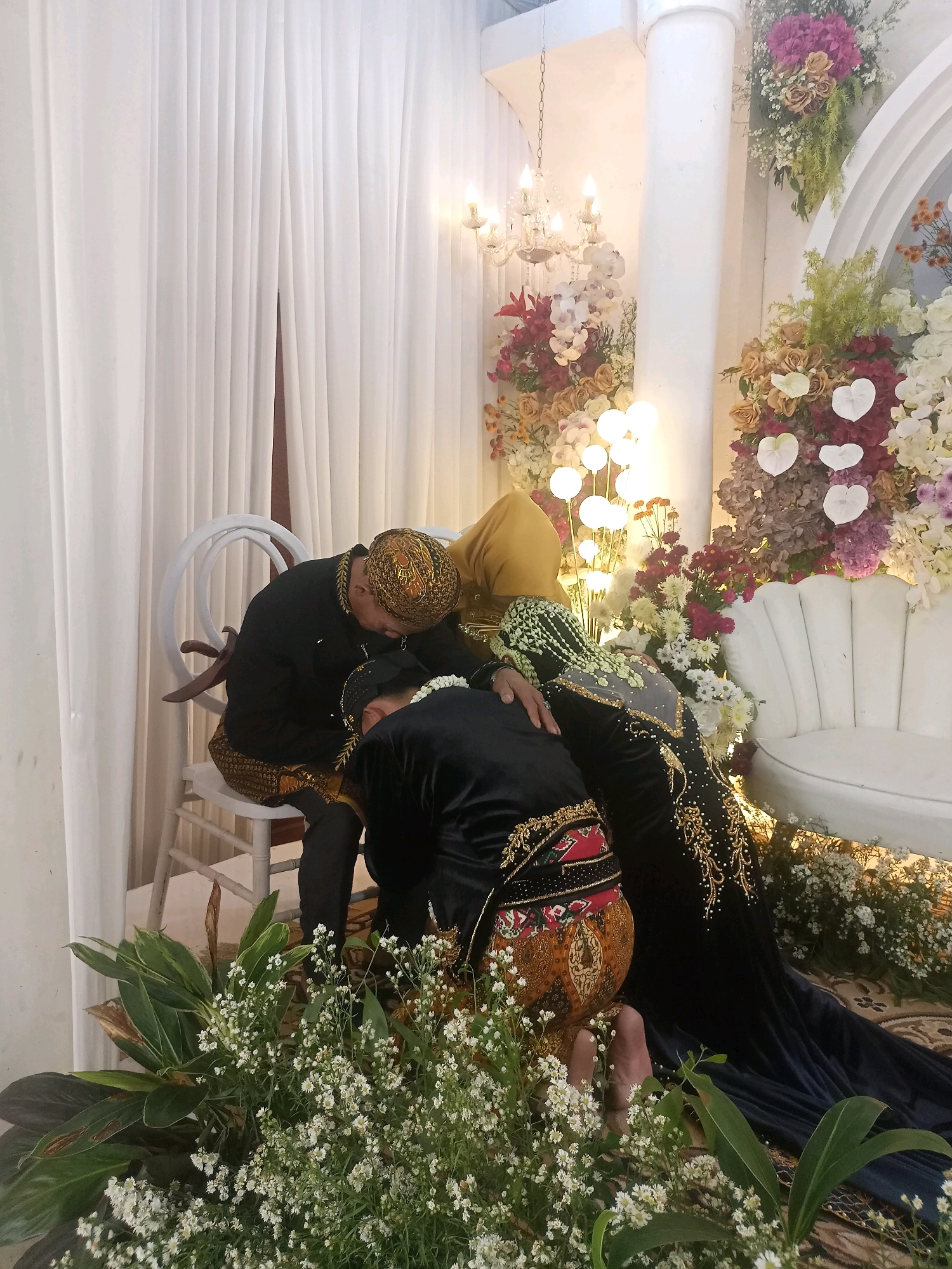 In an indoor wedding venue in Badamita, Indonesia, two individuals in traditional Indonesian wedding attire are engaged in a customary ritual. One person, adorned in a black outfit with gold embroidery and a traditional patterned sarong, is seated on a white chair. The other individual, wearing a black dress with gold embellishments and a yellow headscarf, kneels before the seated person, with their forehead resting on the seated person's knee. A third person, also in a black outfit with gold embroidery and a patterned sarong, is kneeling and appears to be kissing or embracing the seated individual's knee. The environment is decorated with white drapes, floral arrangements featuring various colors of flowers and greenery, a white sofa, and a chandelier. The lighting suggests an evening or indoor event.