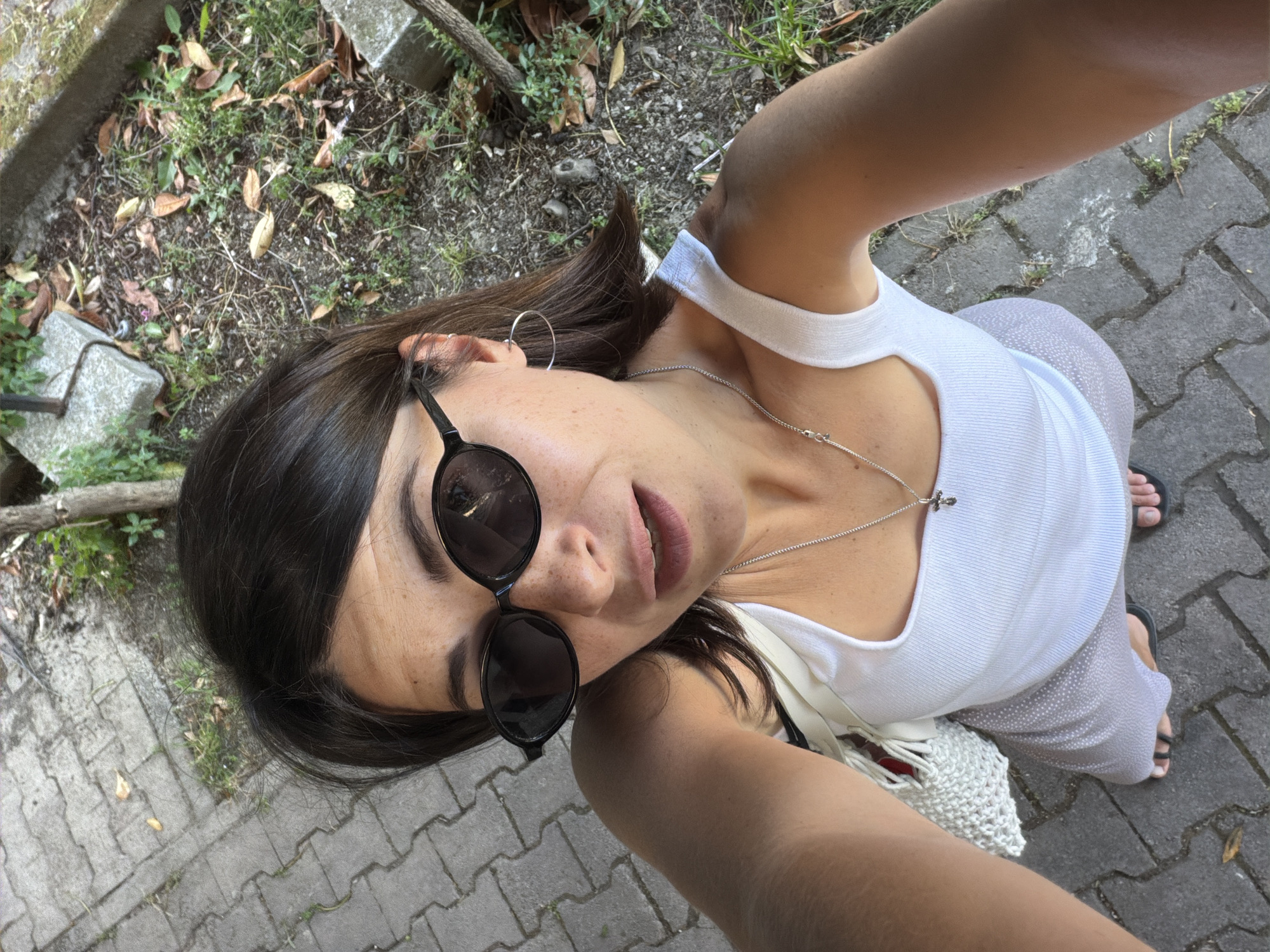 A woman is taking a selfie outdoors in Budva, Montenegro. She wears a white tank top, light patterned pants, and black sunglasses. She has dark hair and is wearing a silver necklace and hoop earrings. The ground is paved with grey bricks. To the left, there is a small patch of dirt with some foliage and debris.