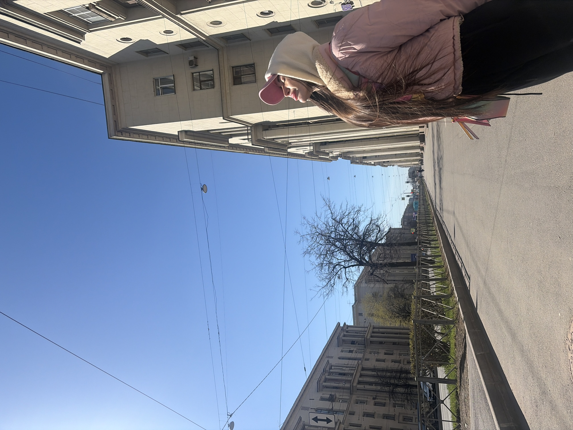 A clear, sunny daytime scene captures a person standing or walking on a city sidewalk, with buildings lining both sides of a wide street, typical of an urban environment such as Saint Petersburg, Russia. The perspective is slightly elevated, looking down the length of the street.
In the foreground, on the right side of the frame, is a person, likely female, viewed from the side and slightly from behind. She is wearing a light pink puffer jacket over a white hooded garment, possibly with a furry or shearling lining, and a pink baseball cap. Her long brown hair flows down her back, and she has colorful, iridescent ribbons or strands woven into it. She appears to be looking downwards or forwards.
The street, made of asphalt, stretches into the distance, with a sidewalk and a low metal fence separating it from a strip of grass and bare trees on the left. On the right, beyond the person, another section of the street is visible. Tall, light-colored stone or concrete buildings with numerous windows and architectural details line both sides of the street, indicating a well-established urban setting. Many overhead utility wires crisscross the bright, cloudless blue sky.
Notable details include the clear, vibrant blue sky suggesting good weather. A bare tree, indicative of early spring or late autumn, stands on the left. A black and white sign with two horizontal arrows pointing in opposite directions is visible on a pole towards the middle-ground. No specific activity beyond being present on the street is discernable. There is no other legible text in the image.