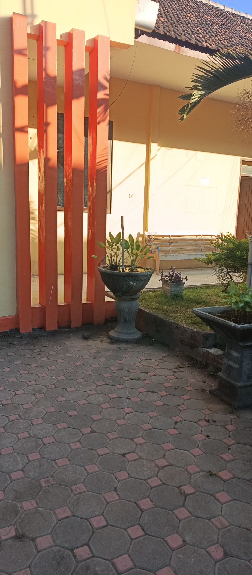 The image depicts an outdoor scene in front of a building, likely a residential or institutional structure, with a tiled walkway. The walkway is paved with hexagonal concrete tiles interspersed with smaller square red tiles, forming a geometric pattern.
In the foreground, to the left, are several vertical orange-colored pillars with a textured surface, casting shadows on the wall behind them. A window is visible between the second and third pillars from the left. To the right of the pillars, there is a large, dark gray, ornate planter with a wide rim, containing several green aquatic plants. The planter rests on a stone pedestal.
Further to the right, a small patch of grass is visible, bordered by a low concrete edge. On the grass, there is a smaller rectangular planter with dark purple foliage. Beyond the planters, a wooden bench is partially visible against the building's light-yellow wall. The building has a tiled roof with brown tiles.
A palm frond extends from the upper right portion of the frame, casting shadows. The scene is illuminated by natural sunlight, creating strong contrasts and shadows. The overall impression is that of a well-maintained outdoor area, possibly a courtyard or a garden adjacent to a building.
Based on the context provided, this scene is located in the city of Arjasa, Indonesia.