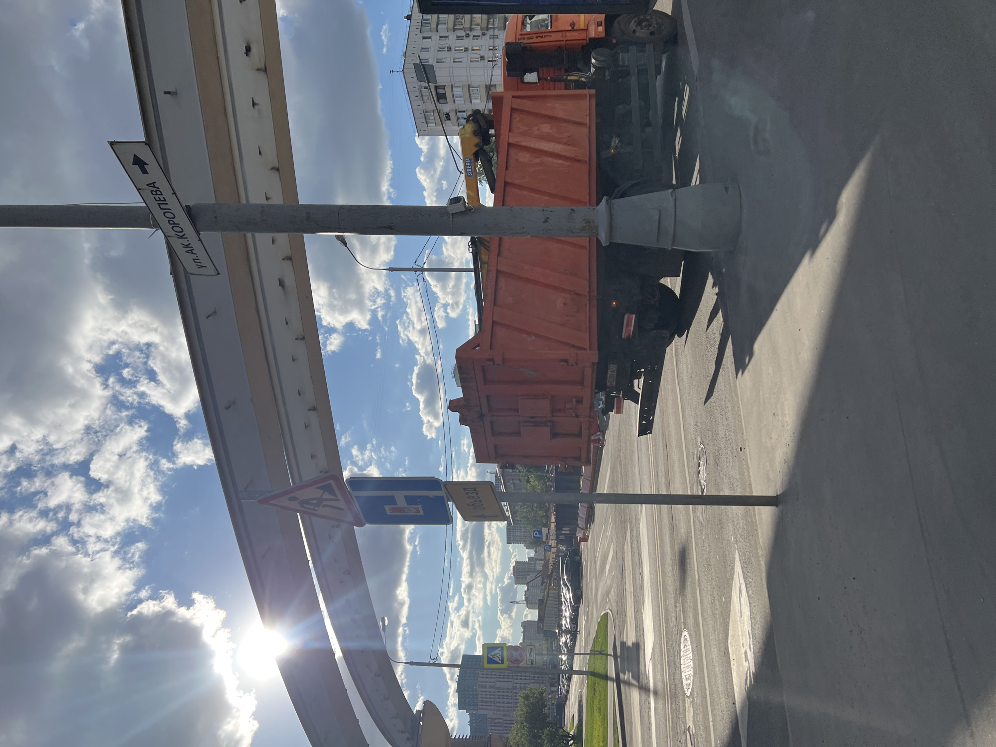 The image shows a street in Khimki, Russia, with roadwork signs indicating construction ahead. A large orange truck with a "Siberia" crane is parked on the street, likely involved in the construction work. A sign on a pole reads "ул.Ак.Королева," indicating the street name is Academician Korolev Street. The street is asphalt with white lane markings and pedestrian crossings. Overpasses are overhead. The sky is blue with scattered clouds, and the sun is shining. Buildings of various sizes and styles are visible in the background, typical of an urban environment. Other cars are on the road. The foreground has a shadow that indicates that the photographer is in the street.