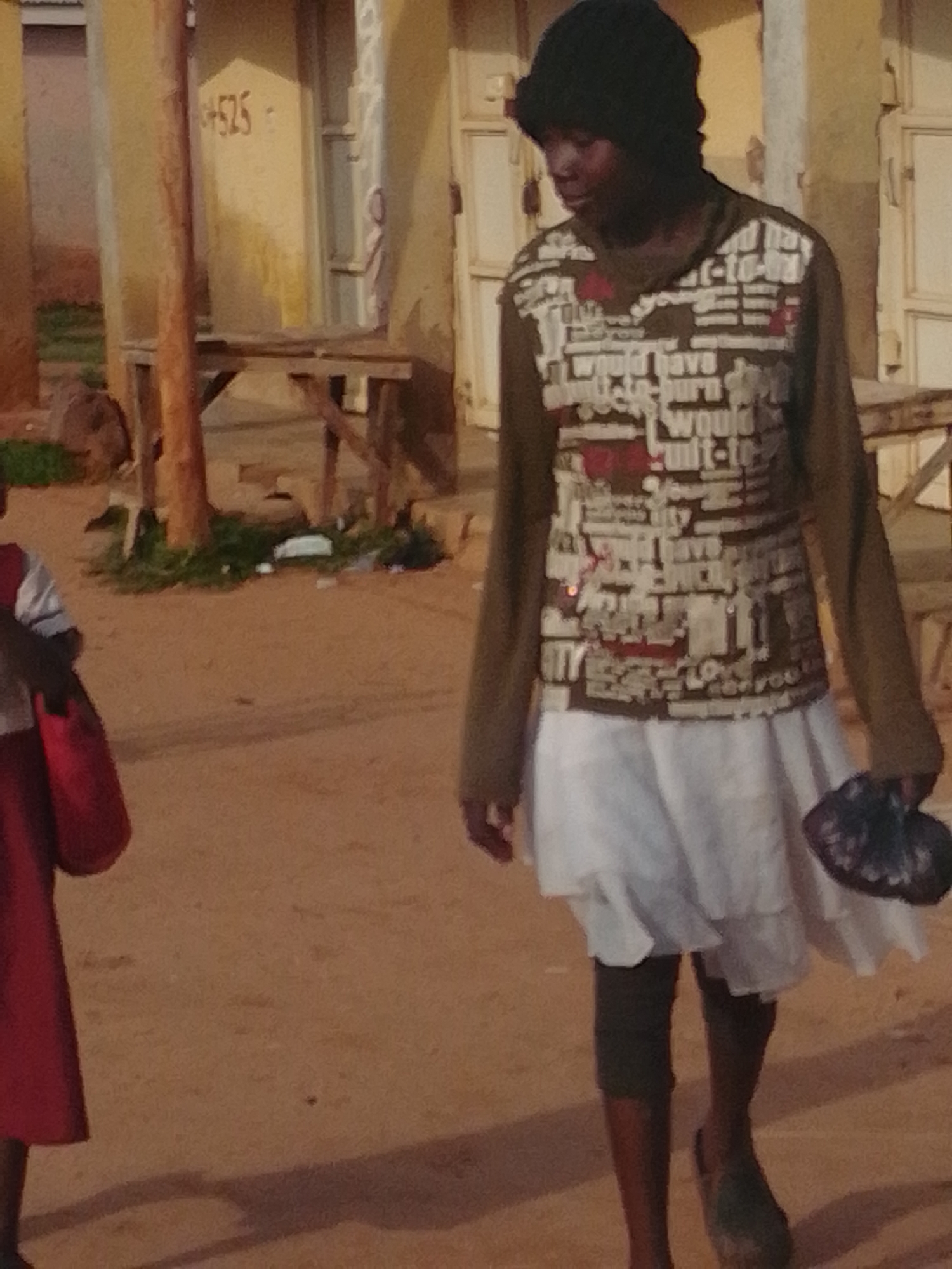 In an outdoor scene in Unknown, Unknown, a young Black individual walks towards the right, carrying a red bag in their left hand and a dark object, possibly a fan or folded item, in their right hand. The individual wears a dark beanie, a long-sleeved shirt with a busy graphic print featuring text, and a white, layered skirt. Dark leggings or knee-high socks are visible beneath the skirt, and the individual is wearing dark shoes. To the left of the frame, another person's legs and a portion of a red garment are visible. The ground is dusty and sandy. In the background, a wooden structure is present, with a building behind it that has yellow walls and visible doorways or windows. A number "4525" is painted on the yellow wall of the building. Green vegetation and scattered debris are also visible on the ground.