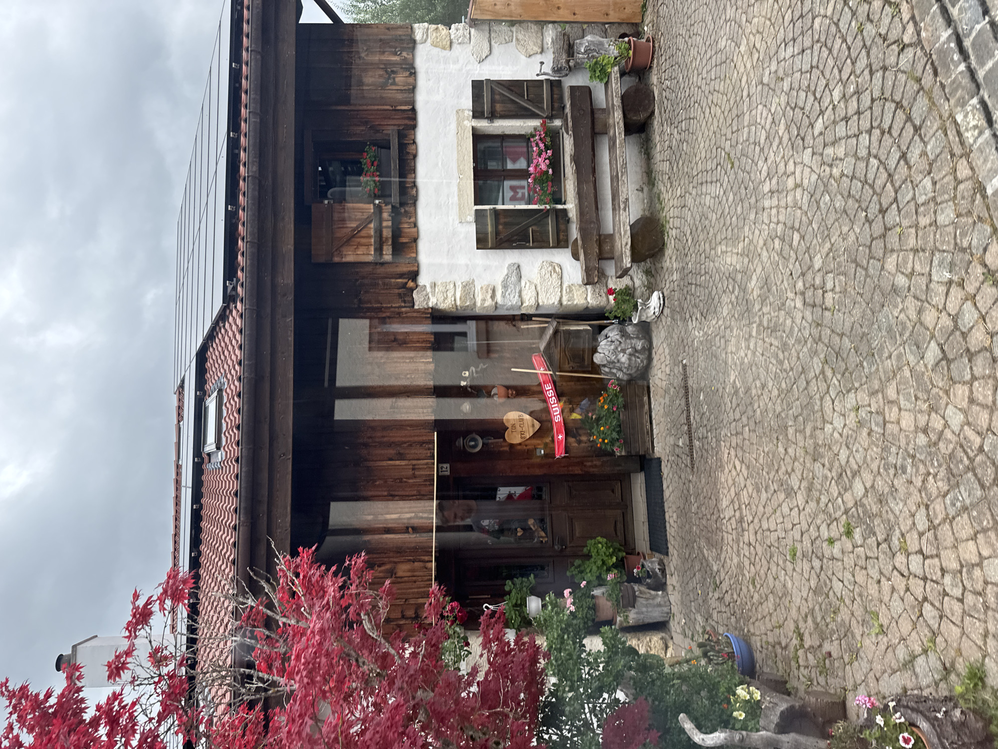 The image depicts a two-story building with a rustic facade, situated on a cobblestone courtyard. The lower story features stone construction, while the upper story is clad in dark wood. Solar panels are installed on the red-tiled roof. A bright red Japanese maple tree stands to the left of the entrance, its vibrant foliage contrasting with the more subdued tones of the building.
The entrance to the building is a double wooden door with a glass pane, through which a person can be seen. Above the door, the number "12" is displayed. To the right of the entrance, there is a wooden sign shaped like a heart with text that appears to read "TON SKI-CLUB". A red banner with "SUISSE" written in white lettering is also displayed to the right of the door. Several potted plants with pink and orange flowers are placed around the entrance and on the facade.
A wooden bench with a rough-hewn appearance is positioned in front of a window on the lower story, which has dark wooden shutters. This window has a flower box filled with pink flowers. Another window above it also has wooden shutters and a dark wooden frame.
The cobblestone courtyard extends from the foreground to the building. Various decorative elements are scattered around the courtyard, including a pair of wooden logs supporting the bench, a small wooden structure resembling a mailbox, a white boot, and several potted plants. The environment suggests a rural or semi-rural setting in Petit-Val, Switzerland, likely a residential or commercial property with a strong connection to local traditions. The sky is overcast, indicating cloudy weather.