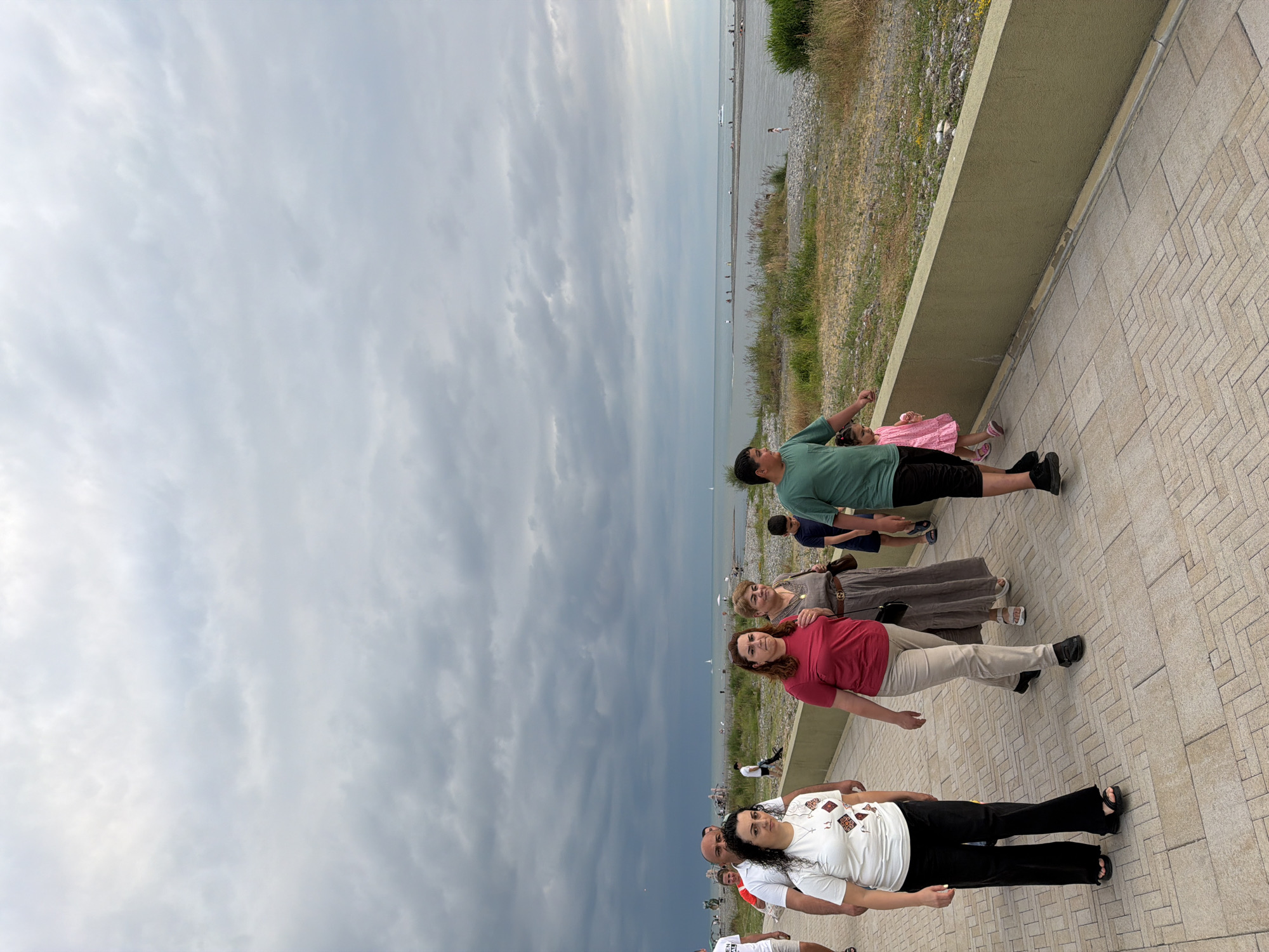 The image shows a group of people standing on a paved walkway next to a body of water, likely the sea. The sky is overcast with dramatic clouds, suggesting a dramatic weather change. The people are dressed casually, indicating a relaxed outdoor setting. Some are looking towards the water, while others are looking towards the camera.
In the foreground, a woman in a white shirt and black pants is prominently featured. Behind her, a man and woman appear to be in conversation. Further back, a group of people, including adults and children, are standing together. One child in a pink dress is visible. The background shows a beach with a few people scattered along it and the sea stretching out to the horizon. The overall scene suggests a gathering of people enjoying a day out by the water, despite the cloudy sky. The location appears to be a promenade or boardwalk.
The time of day is difficult to determine precisely due to the overcast conditions, but the light suggests it is daytime. The weather is cloudy, with the clouds creating a somber mood in the sky, contrasting with the potentially pleasant activity of the people. There is no visible text in the image. The paved walkway has a patterned brick design. The greenery along the edge of the walkway adds a natural element to the urban setting.