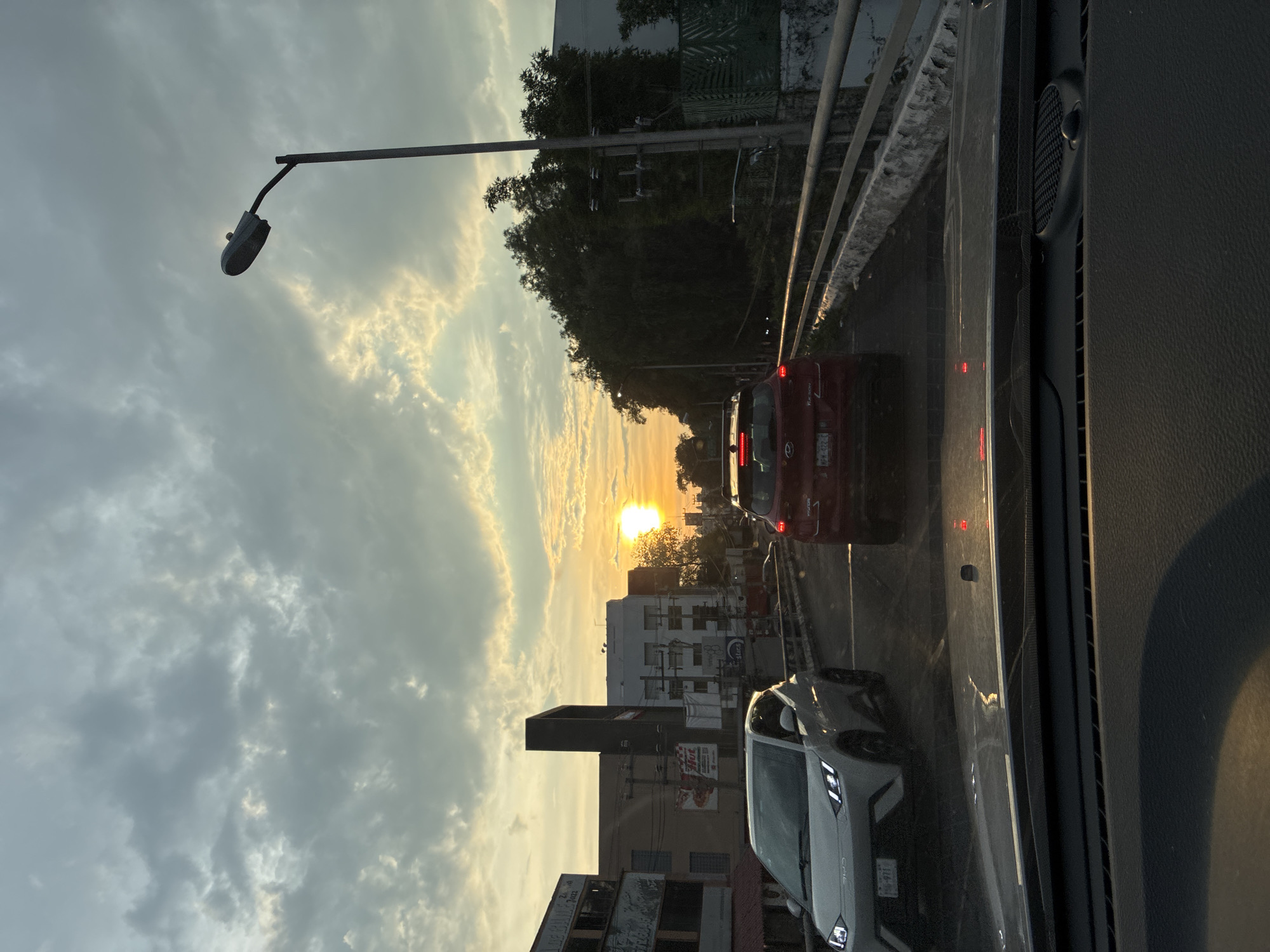 View from inside a vehicle moving along a city street in Naucalpan de Juárez, Mexico, as the sun sets. A streetlight is on the left, and traffic includes a red car and a white car ahead. Buildings line the street. The sky is filled with clouds.