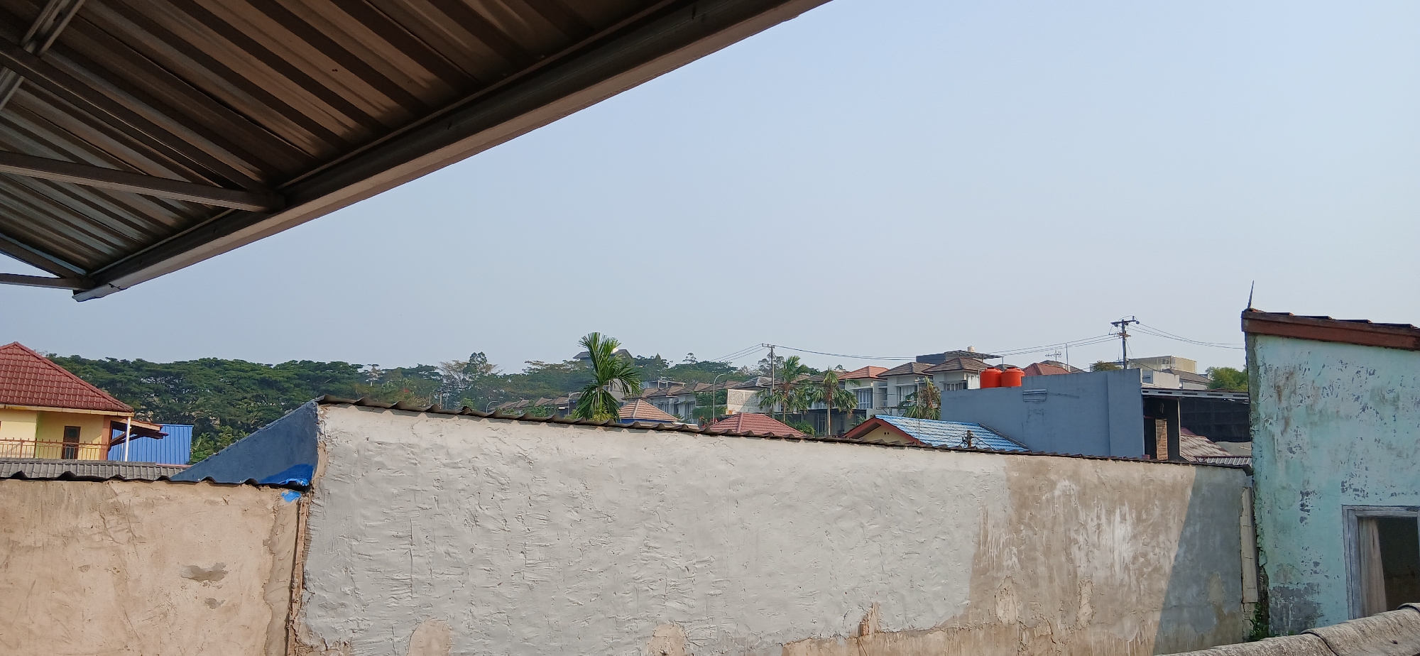 The image shows a view of a residential area with houses and buildings under a hazy sky. In the foreground, a textured, off-white concrete wall runs horizontally across the frame, with a portion of a corrugated metal roof extending from the top left corner. Behind this wall, the roofs of several houses are visible, with tiled roofs in various colors including red and brown. A palm tree stands out among the buildings. In the middle ground, more houses are clustered together, some with blue or grey walls and orange water tanks on top of a grey structure. Power lines and a utility pole are also visible. The background consists of a dense green treeline, suggesting a forested area or large trees surrounding the neighborhood. The overall impression is that of an urban or suburban environment on a clear but slightly overcast or hazy day. The scene is located in Samarinda, Indonesia. There are no people visible in the image.