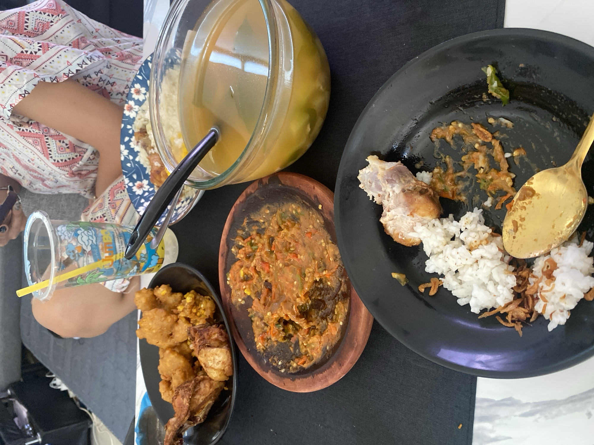A overhead shot captures a table setting with a meal in progress. In the foreground, a black plate holds remnants of a meal, including white rice, a piece of fried chicken, and a dollop of red chili paste. A golden spoon rests on the plate. Adjacent to this, a small, rustic wooden dish contains more of the red chili paste, appearing to be a condiment. Further back, a clear glass bowl is filled with a yellowish broth, likely a soup, with some green vegetables visible within. Another plate, decorated with blue floral patterns, sits behind the soup, holding a piece of fried chicken and rice. To the left, a black bowl contains a serving of fried chicken pieces. A clear plastic cup with a colorful cartoon design and a yellow straw is also present, suggesting a beverage. The table is covered with a black tablecloth, with a portion of a white and patterned garment, likely worn by a person seated at the table, visible at the top. The background shows a blurred view of a gray sofa, indicating an indoor setting. The presence of Indonesian dishes like fried chicken and chili paste strongly suggests the location is in Indonesia, potentially in a casual dining setting in Cibinong, a city known for its culinary offerings.