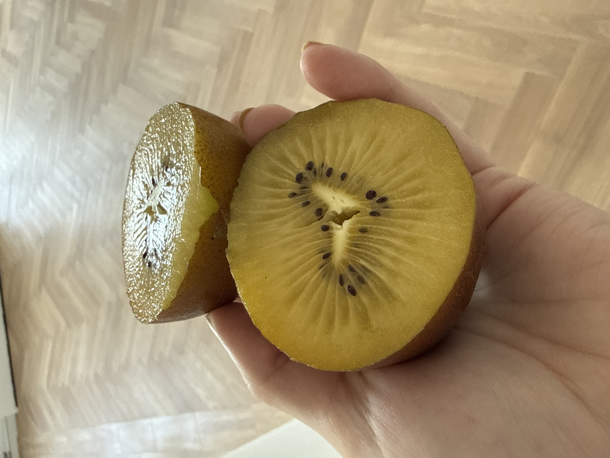 A hand holds two halves of a golden kiwi fruit against a background of wood-patterned flooring. The kiwi fruit has a golden flesh with small, black seeds arranged in a radial pattern. The hand appears to be light-skinned. The flooring suggests an indoor setting. No specific information connects the image to Moscow, Russia.