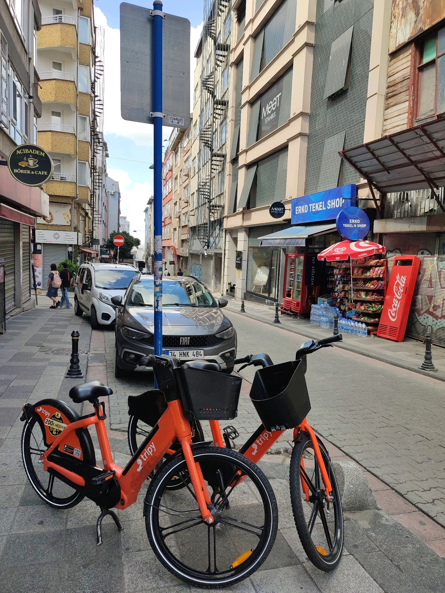 This is a street-level, eye-level, outdoor, daytime, medium shot photograph of a street scene in Kadıköy, Turkey. The foreground features two orange shared electric bicycles. One bicycle is closer to the camera, showing its front wheel, handlebars, and basket. The second bicycle is partially visible behind it.
The street is paved with grey cobblestones. In the mid-ground, a grey Fiat car with license plate "34 HNK 484" is parked on the right side of the street. To the left of the Fiat, a white van is also parked. A black bollard stands in front of the Fiat. Two figures, one with a backpack, are walking away from the camera on the left side of the sidewalk.
The background shows a narrow street lined with buildings on both sides. The buildings have multiple floors with spiral fire escapes on their exteriors. On the right side, a shop with a blue sign reading "ERKO TEKEL SHOP" is visible. In front of this shop, there is a display of bottled water, a red Coca-Cola branded cooler, and a red umbrella shading shelves stocked with various items. Signs for "MEET ISTANBUL HOTEL" and "ACIBADEM BÖREK & CAFE" are also present on the buildings. A blue and grey pole with a sign is positioned in the center of the scene. The sky above is partly cloudy.