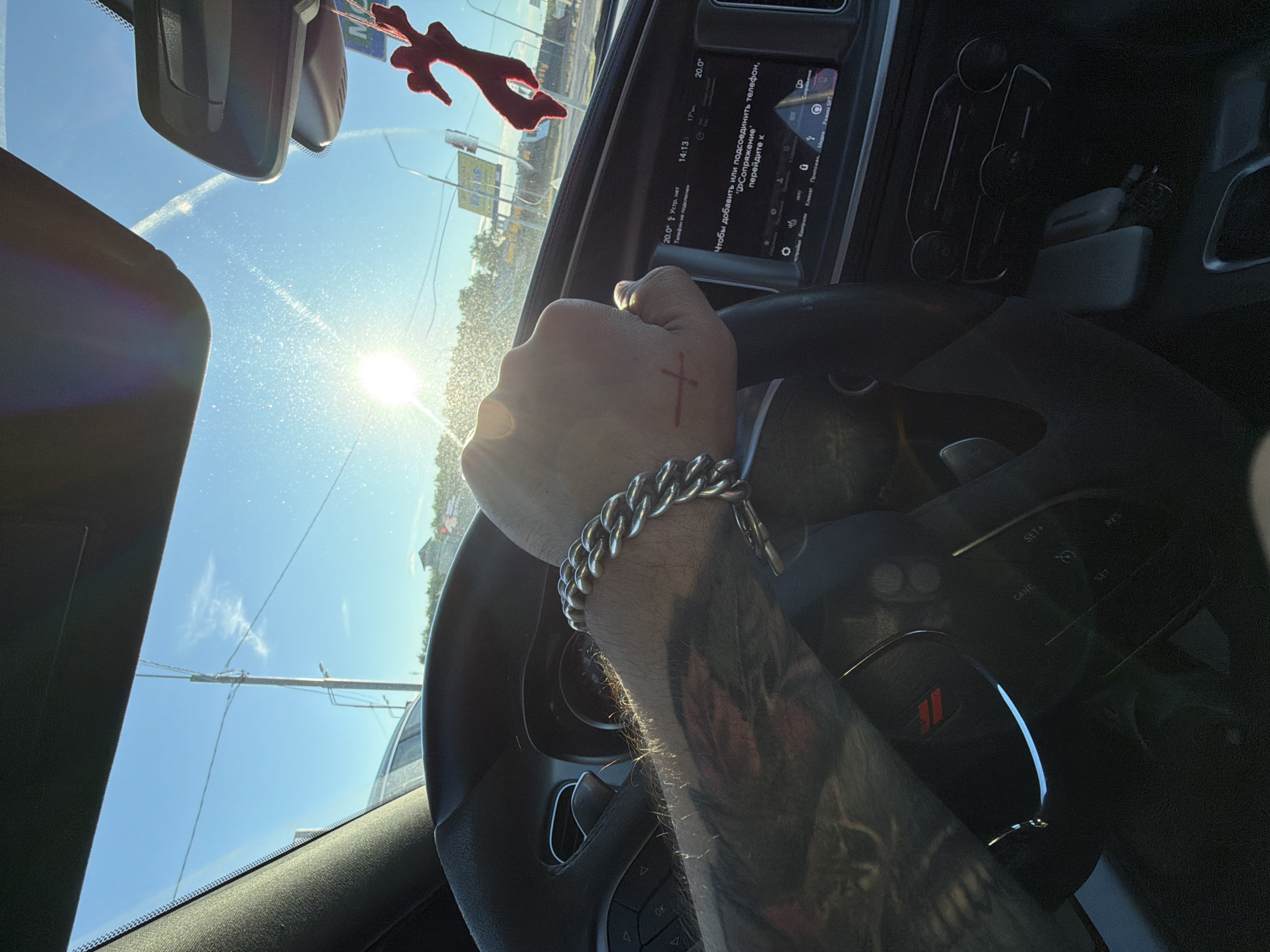The image shows a first-person perspective from inside a car, likely in Pokrovskoe, Russia, on a sunny day. The driver's hand, adorned with a thick silver bracelet and a small red cross tattoo on the knuckles, is gripping the steering wheel. The steering wheel has a black leather finish with a red Dodge logo in the center. The car's dashboard features a digital display showing the time as 14:13 and the temperature as 20.0°C. Text on the screen, in Russian, appears to be related to phone connectivity. Outside the car, the bright sun is visible through the windshield, creating lens flare. Power lines and trees are visible in the distance, suggesting a semi-urban or suburban environment. A red, car air freshener shaped like an animal hangs from the rearview mirror. The sky is clear and blue.
