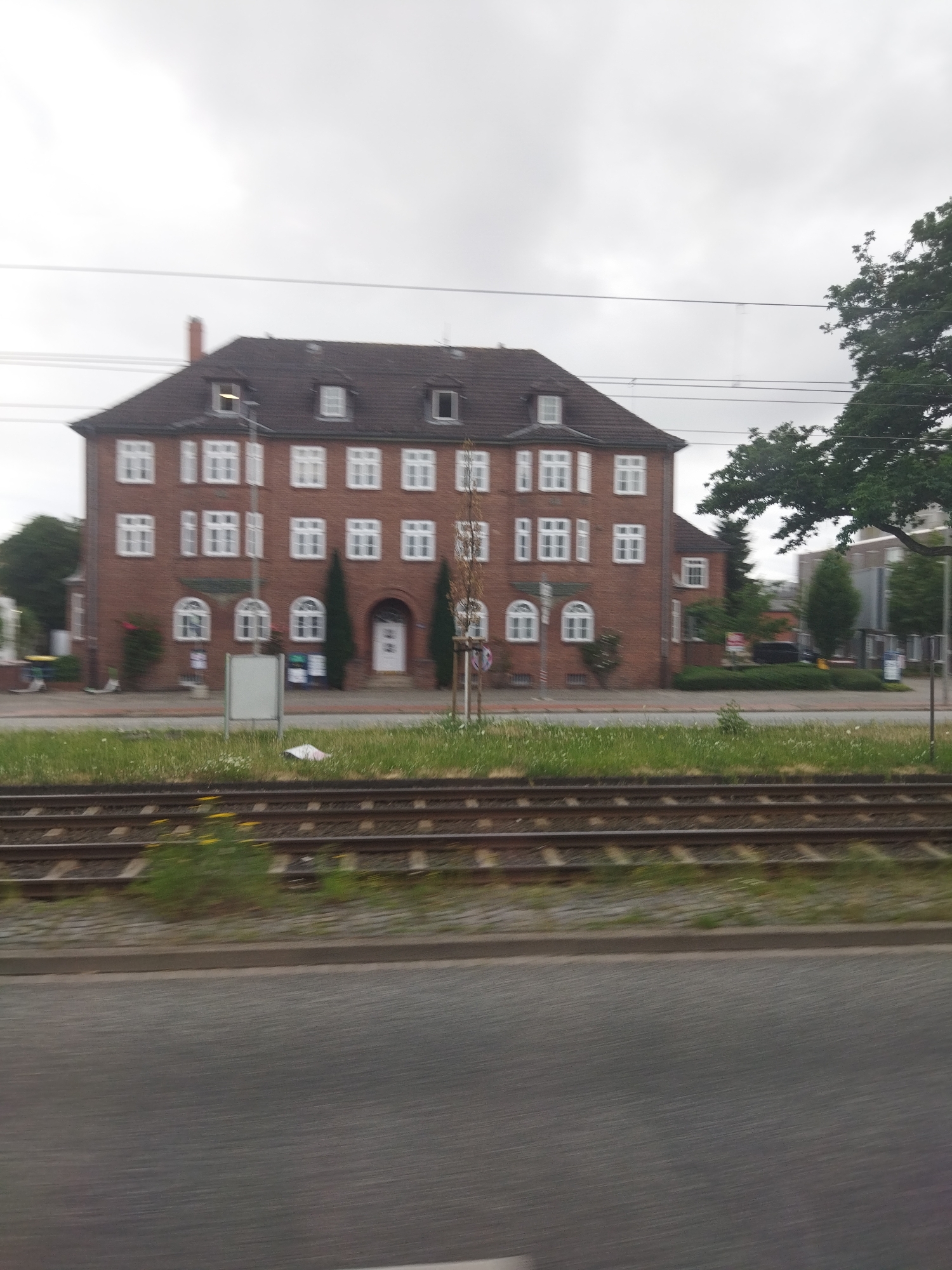 The image captures a three-story brick building with a dark tiled roof and dormer windows, situated in Hanover, Germany. The building has numerous white-framed windows and a prominent arched entryway with a white door. In front of the building, there's a grassy area with some scattered wildflowers, a paved walkway, and a set of train tracks. The sky is overcast, and the overall scene appears to be captured from a moving vehicle, as evidenced by the motion blur on the foreground and the train tracks. Trees are visible on the right side of the building and further in the background. A white rectangular sign or board stands in front of the building, and some smaller signs or posters are attached to the building's facade.