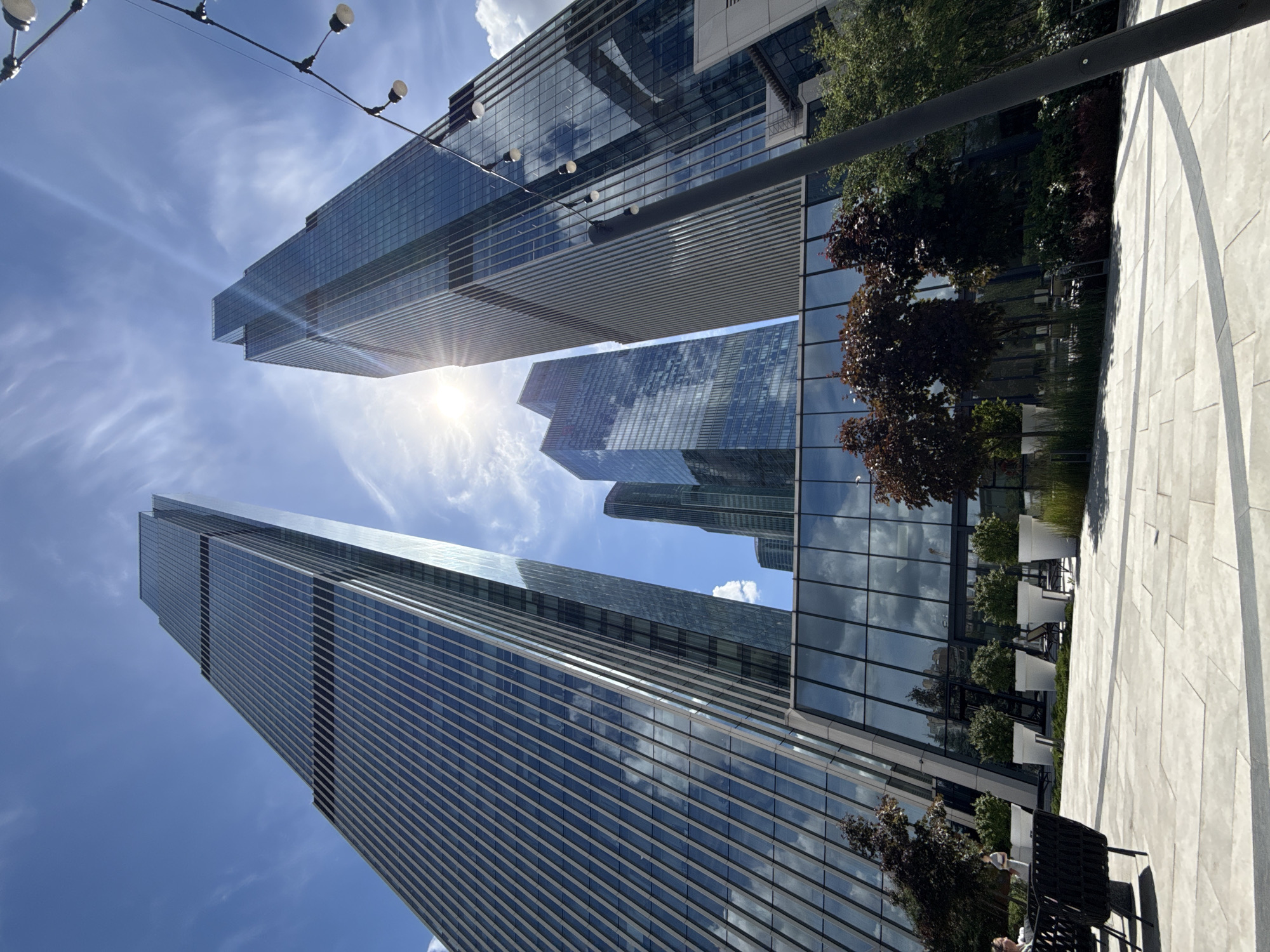 The image depicts a low-angle, wide shot of the Moscow City business district on a bright, sunny day. The foreground features a paved plaza with geometric grey and white tiles, leading to a seating area with dark wicker chairs and tables. Beyond the plaza are several modern glass skyscrapers, their facades reflecting the blue sky and scattered white clouds. The sun is prominently visible, creating lens flare and illuminating the scene.
In the mid-ground, a line of white planter pots containing lush green bushes and small trees is visible, separating the plaza from the buildings. Behind these planters, the lower levels of the skyscrapers showcase large glass windows, also reflecting the sky and surrounding environment. The architecture of the buildings is characterized by sharp angles and sleek, vertical lines. A string of exposed light bulbs hangs from a dark pole on the right side of the frame, adding an element of outdoor ambiance.
In the background, the full height of the skyscrapers is apparent, with the tallest structures dominating the skyline. Some of these buildings have visible horizontal banding or patterns in their glass exteriors. The sky is a vibrant blue, with wispy white clouds stretching across it. A few individuals are sparsely visible in the distance on the plaza, suggesting a public outdoor space. The overall scene conveys a sense of modern urban development and contemporary city life in Moscow.