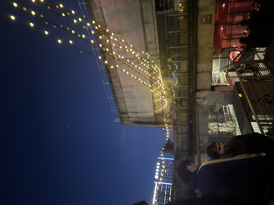 The image captures an outdoor urban scene at dusk or night, illuminated by numerous warm yellow string lights and distant city lights, under a deep blue sky.
In the upper portion of the frame, the dark blue sky is prominent, featuring a distinct, thin crescent moon and a few faint stars. Several strings of bright yellow fairy lights crisscross the upper sky, stretching horizontally.
Below these lights, a robust concrete structure, possibly a bridge, overpass, or the underside of a building's upper level, extends across the middle of the image. More strands of the same yellow lights are draped beneath this concrete structure, creating a festive canopy effect. To the right of this structure, on a vertical wall, a pixelated artwork resembling a classic "Space Invaders" character is visible, suggesting urban art or graffiti.
Further to the right, behind metal railings, an area is bathed in a reddish glow, likely from internal lighting or a neon sign of a commercial establishment. Silhouetted figures of people can be seen gathered in this red-lit space.
The lower part of the image features a gathering of people in the foreground, mostly seen from behind or in profile. One person with dark, curly hair is prominent on the right. Another individual with shorter dark hair and a beard is visible further left. Their presence suggests a social gathering, possibly at an outdoor bar, restaurant patio, or public square.
In the distant background, to the left, a modern high-rise building stands tall, its windows brightly illuminated with various white and blue lights, contributing to a vibrant city skyline.
The overall atmosphere is one of a clear, mild evening, with people enjoying the urban ambiance and decorative lighting. No specific text is clearly discernible in the image.