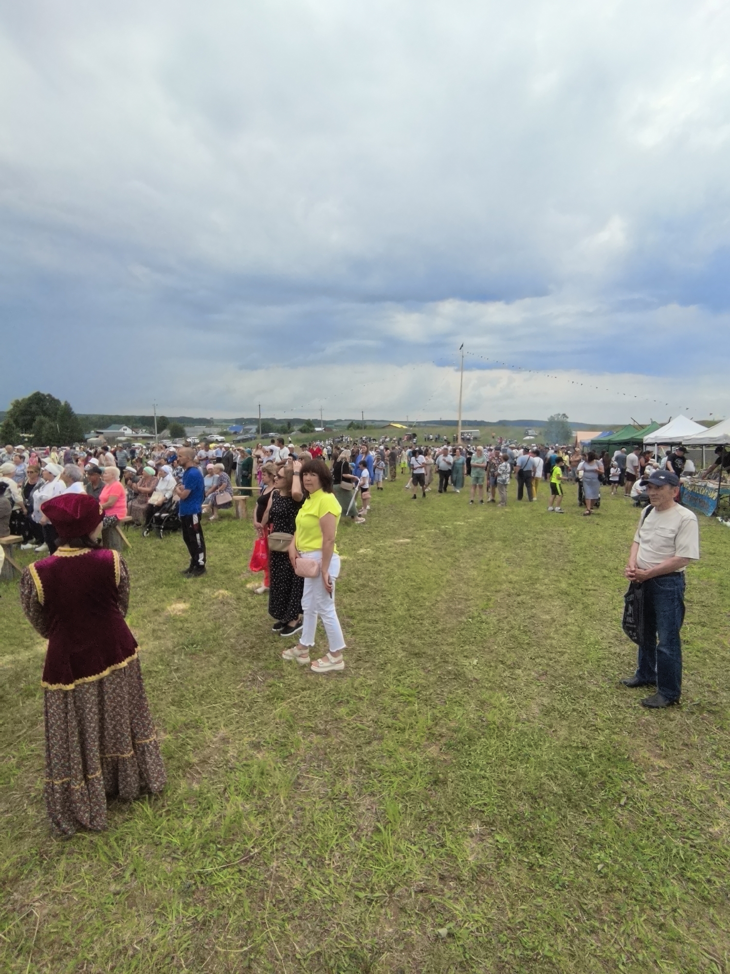 This image depicts an outdoor event, likely a festival or gathering, on a grassy field in the village of Koyanovo, Russia. A large crowd of people, consisting of men, women, and children, are present. Many are standing and observing, while some are seated on benches in the background. Several tents and stalls are visible in the middle ground, suggesting vendors or information booths. In the foreground, a person in traditional clothing stands with their back to the camera, facing the crowd. Another man in a light-colored shirt and jeans stands to the right, looking towards the viewer. The sky is overcast with dramatic clouds. The overall atmosphere appears to be that of a community event.