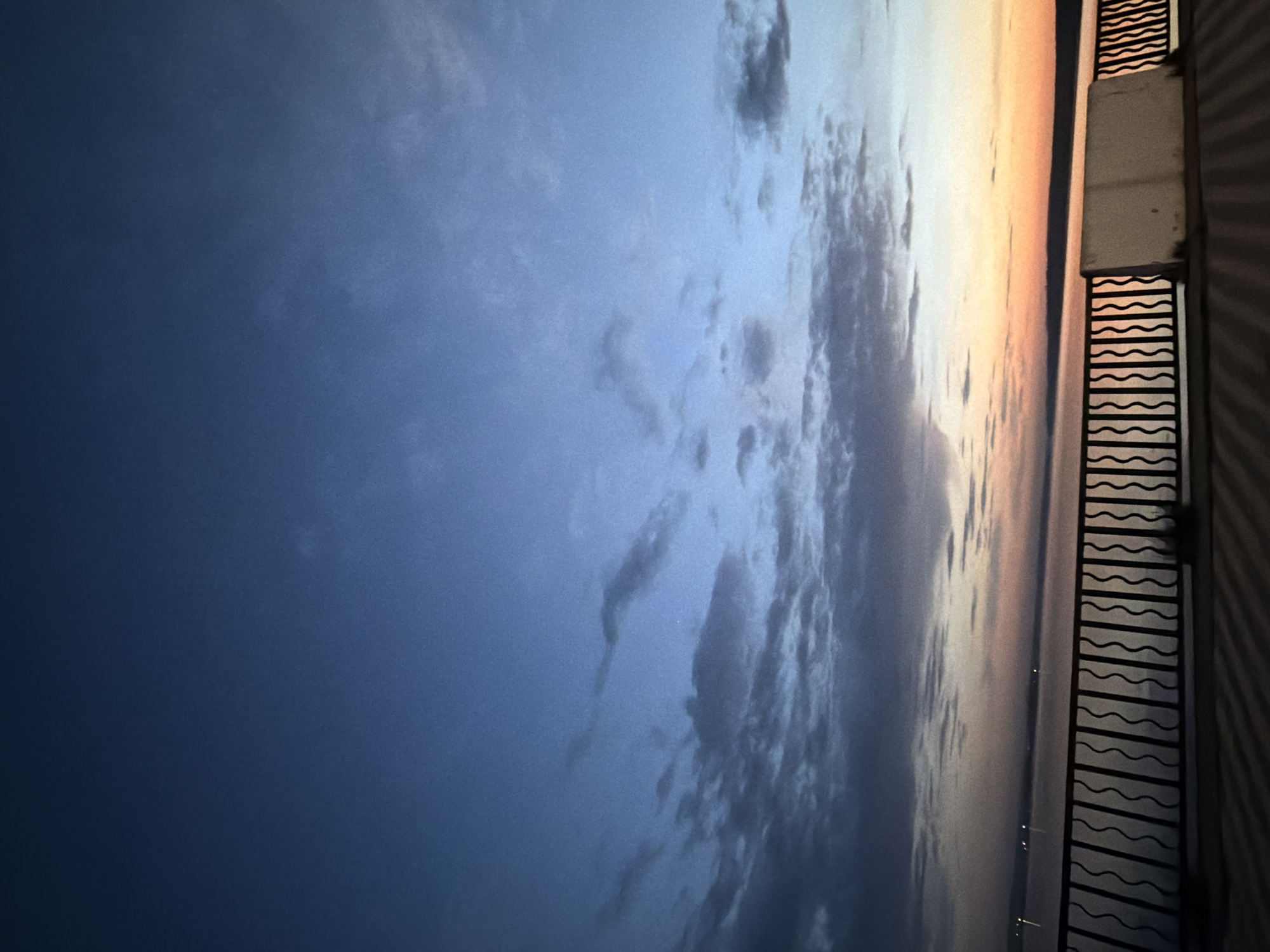 Here is a detailed description of the image:
**Scene:** The image shows a dusk or dawn scene from a balcony. The sky is prominently featured, transitioning from a deep blue on the left to lighter hues with hints of orange and peach on the right. Clouds are present in the center, stretching vertically.
**Objects:** A dark-colored balcony railing with a wavy design runs along the right edge of the frame. A concrete or light-colored structure is visible behind the railing. The suggestion of water is visible in the middle right, with light sources appearing to be on the water.
**Environment:** The setting appears to be urban, with the city lights scattered along the horizon. The scene evokes a sense of quiet observation from a high vantage point.
**Location:** The city and specific location within are unknown.