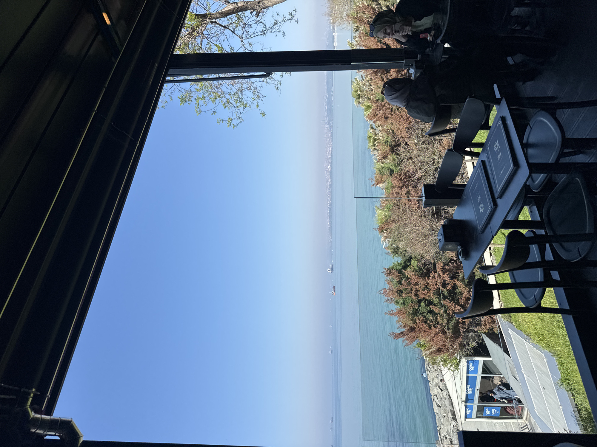 The image captures a view from an elevated indoor space, possibly a restaurant or cafe, overlooking the Marmara Sea in Bakırköy, Turkey. In the foreground, a black table with two menus labeled "1864" is surrounded by black chairs. Two individuals are seated at the table; one is using a phone.
Beyond the immediate dining area, the Marmara Sea stretches to the horizon, dotted with ships. A city skyline is visible in the distance, across the water. The shoreline features vegetation, including trees and shrubbery.
Visible on the right is a partial view of the exterior of the building including a sign in a blue color. The image is framed by architectural elements of the building, including roof. The sky is clear and blue, indicating good weather conditions.