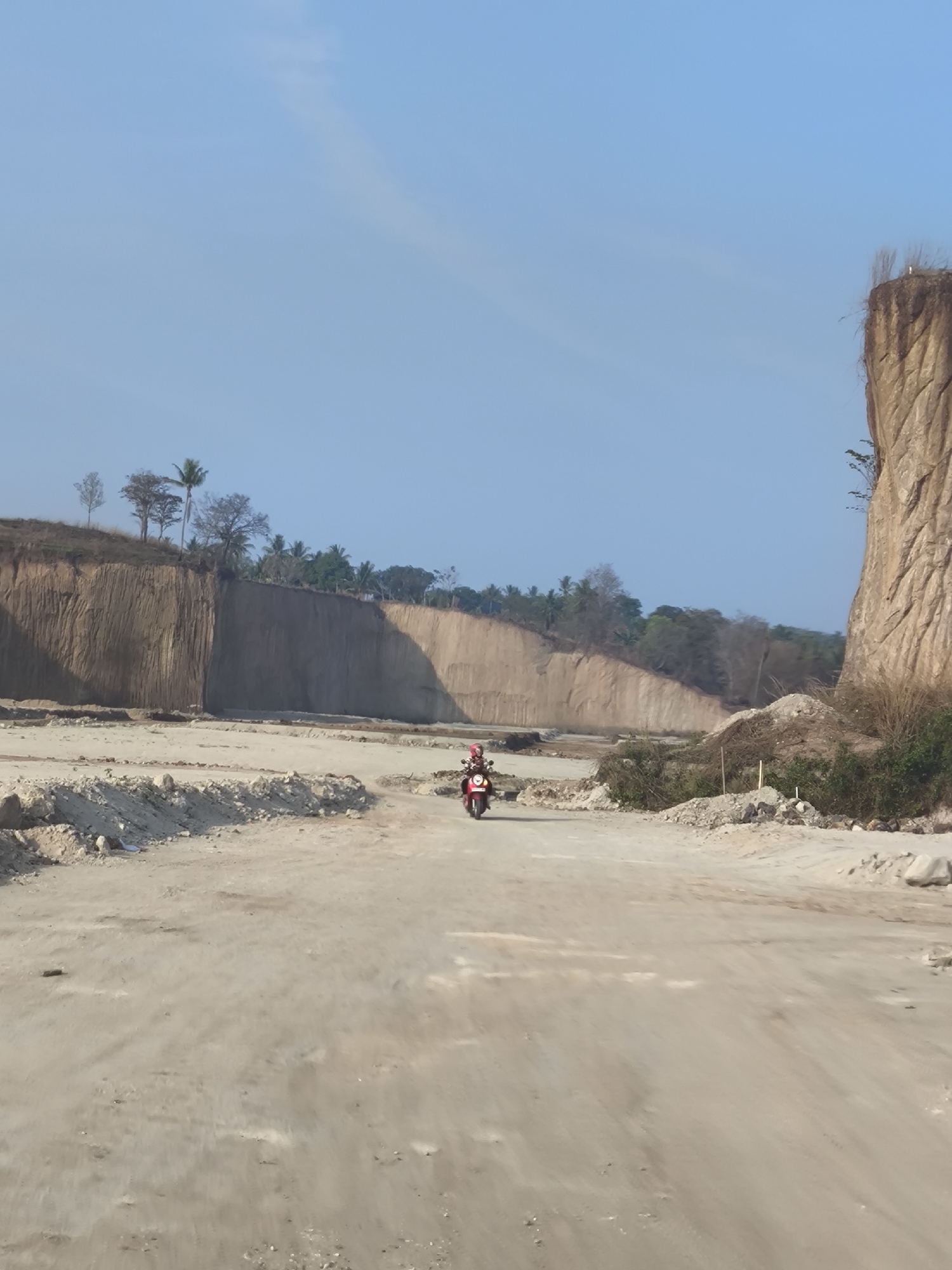 A person rides a red motorbike down a dusty, unpaved road in a quarry. Steep, eroded cliffs of tan earth rise behind the road, with trees and vegetation along the tops and edges. The sky is a clear, pale blue. The scene appears to be in a quarry or mining area, possibly part of the industrial landscape of Cilegon, Indonesia, known for its heavy industry and mining operations. The road is rough and appears to be under construction or in a state of disrepair.