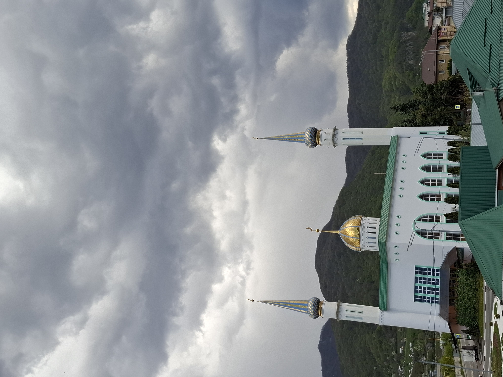 The image shows a white mosque with green roofs and two minarets, one with a golden dome and the other two with blue domes, in Karachayevsk, Russia. The background features a mountain range and a cloudy sky. The mosque is situated near a road and other buildings.